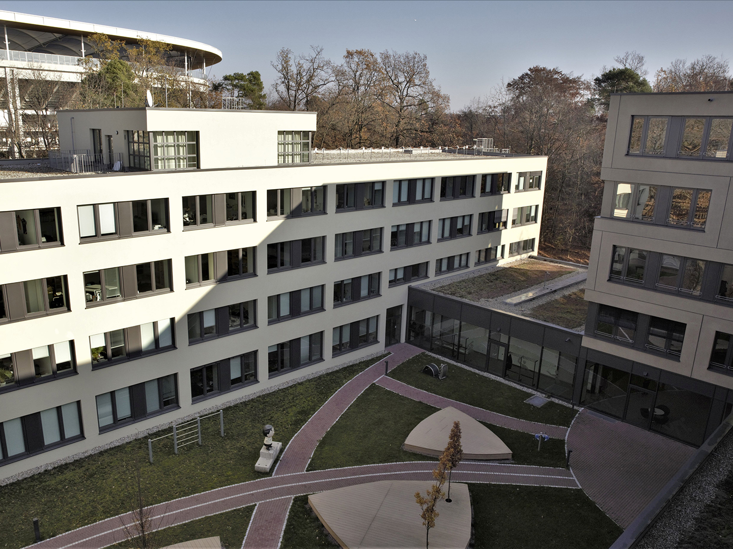 Blick auf einen modernen Gebäudekomplex mit mehreren Etagen, umgeben von einem gepflegten Innenhof mit Wegen und Rasenflächen. Im Hintergrund sind Bäume zu sehen, die auf eine naturnahe Umgebung hinweisen.