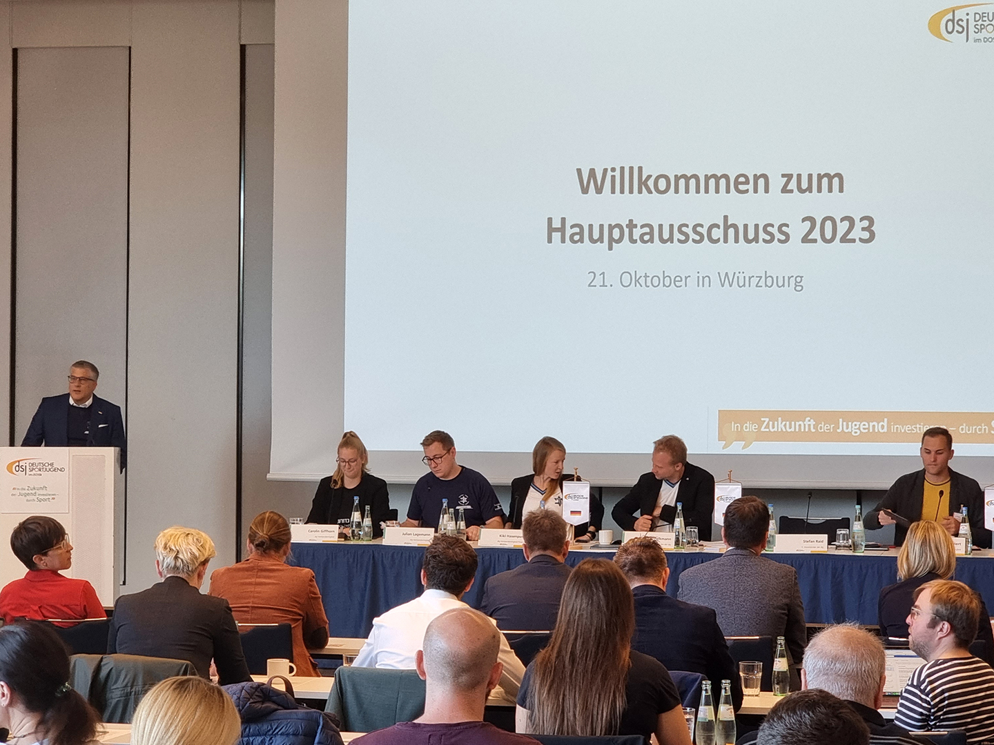 Eine Konferenzsituation mit einem Redner am Pult und mehreren Personen an einem Tisch. Im Hintergrund steht ein Bildschirm mit der Begrüßung zum Hauptausschuss 2023 in Würzburg. Die Anwesenden scheinen aufmerksam zuzuhören.