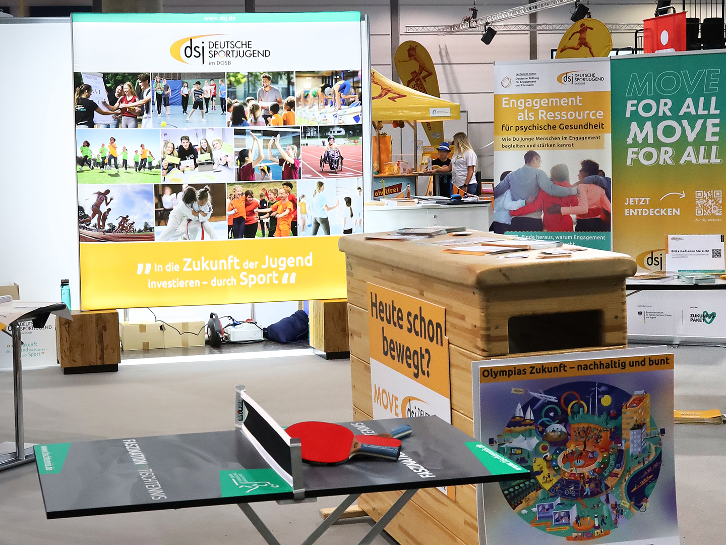 Ein Stand auf einer Veranstaltung mit einem großen Banner der Deutschen Sportjugend im Hintergrund. Im Vordergrund steht ein Tischtennistisch mit einem roten Schläger. Daneben sind Informationsmaterialien und Plakate über sportliche Aktivitäten und Engagement für die Jugend zu sehen.