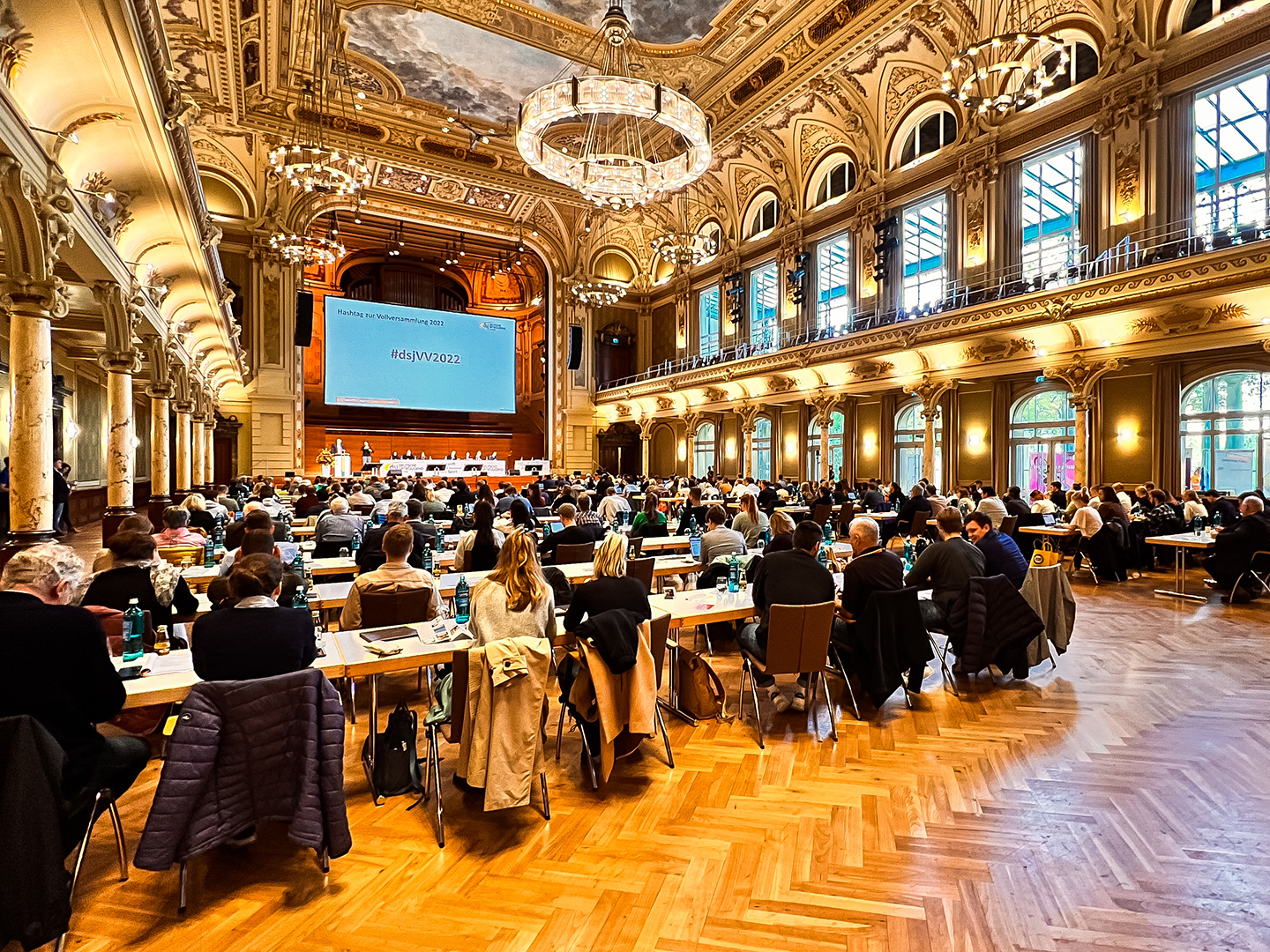 Ein großer Konferenzsaal mit vielen Teilnehmern, die an Tischen sitzen. An der Wand ist ein Bildschirm mit der Aufschrift "#dsjVV2022". Der Raum hat hohe Decken, große Fenster und eine elegante Einrichtung.