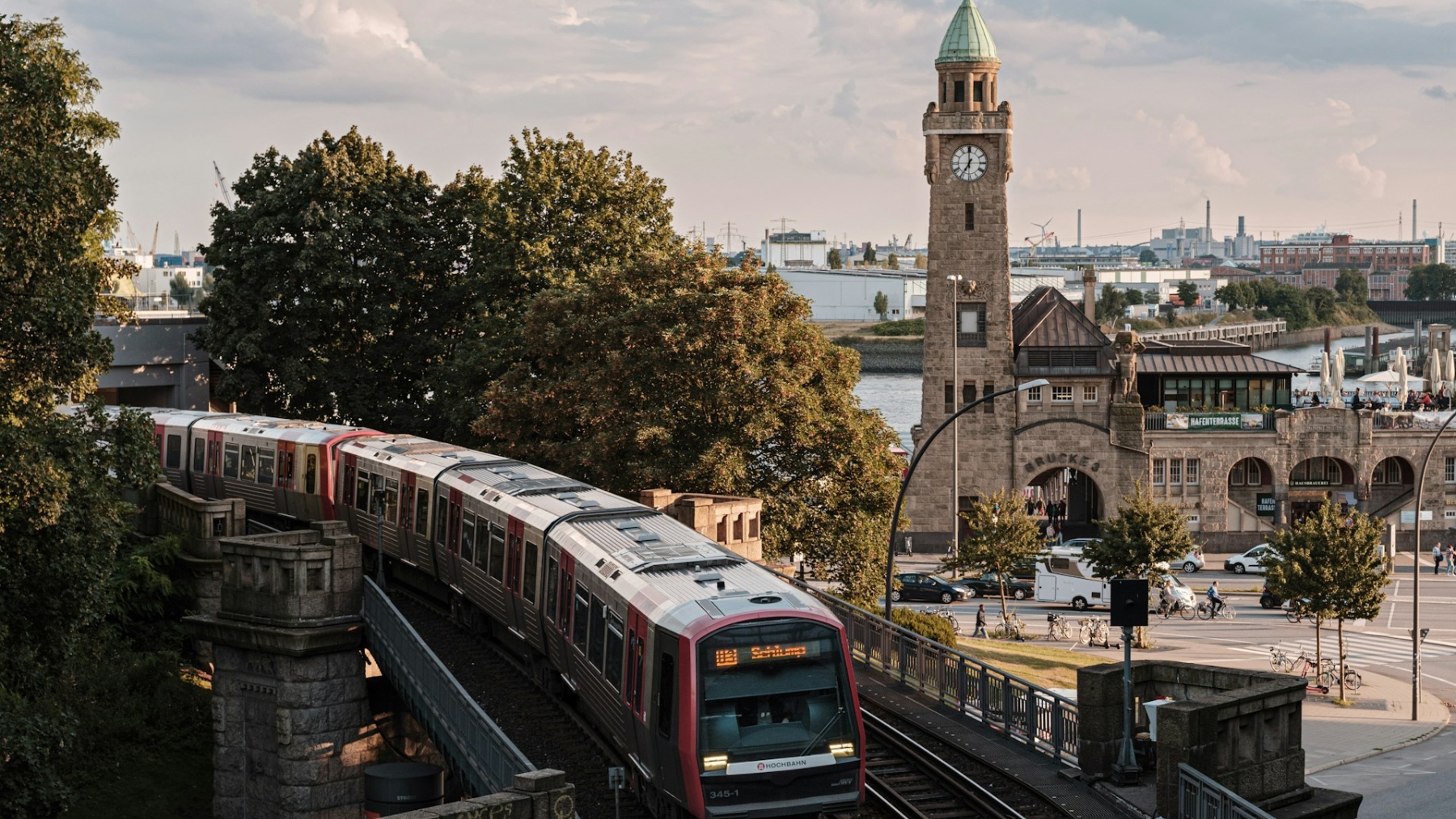 Eine S-Bahn fährt über eine Brücke, während im Hintergrund ein historischer Turm mit Uhr und Bäumen zu sehen ist. Der Himmel ist bewölkt und es gibt eine städtische Umgebung mit Straßen und Fahrzeugen.