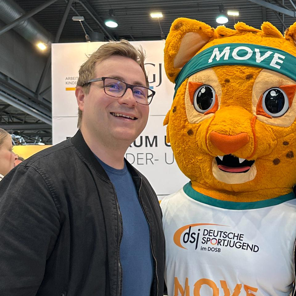 Porträt Julian Lagemann mit Brille lächelt neben einem großen, orangefarbenen Maskottchen, einem Geparden mit einem Stirnband, auf dem "MOVE" steht. Im Hintergrund sind unscharfe Stände einer Veranstaltung zu sehen.