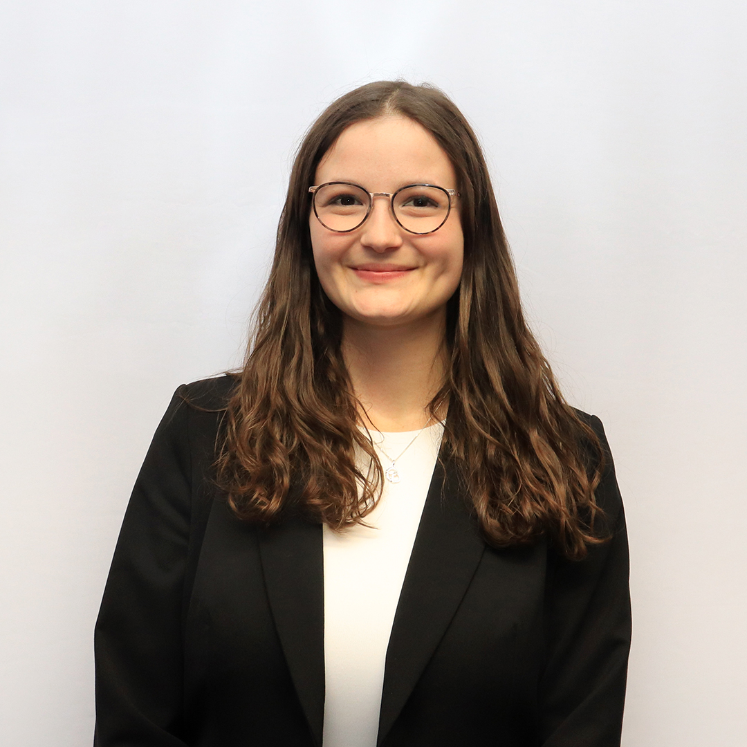 Porträt von Leandra Götz mit langen, lockigen Haaren und Brille trägt einen schwarzen Blazer über einem weißen Oberteil. Der Hintergrund ist hell und neutral.