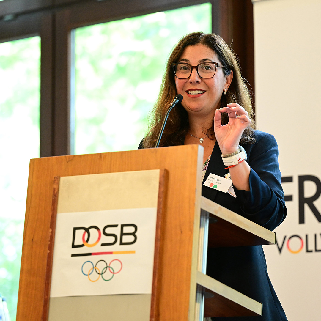 Keren Vogler steht an einem Rednerpult mit dem Logo des DOSB. Sie spricht und hebt eine Hand, während sie lächelt. Im Hintergrund sind Fenster mit natürlichem Licht zu sehen.