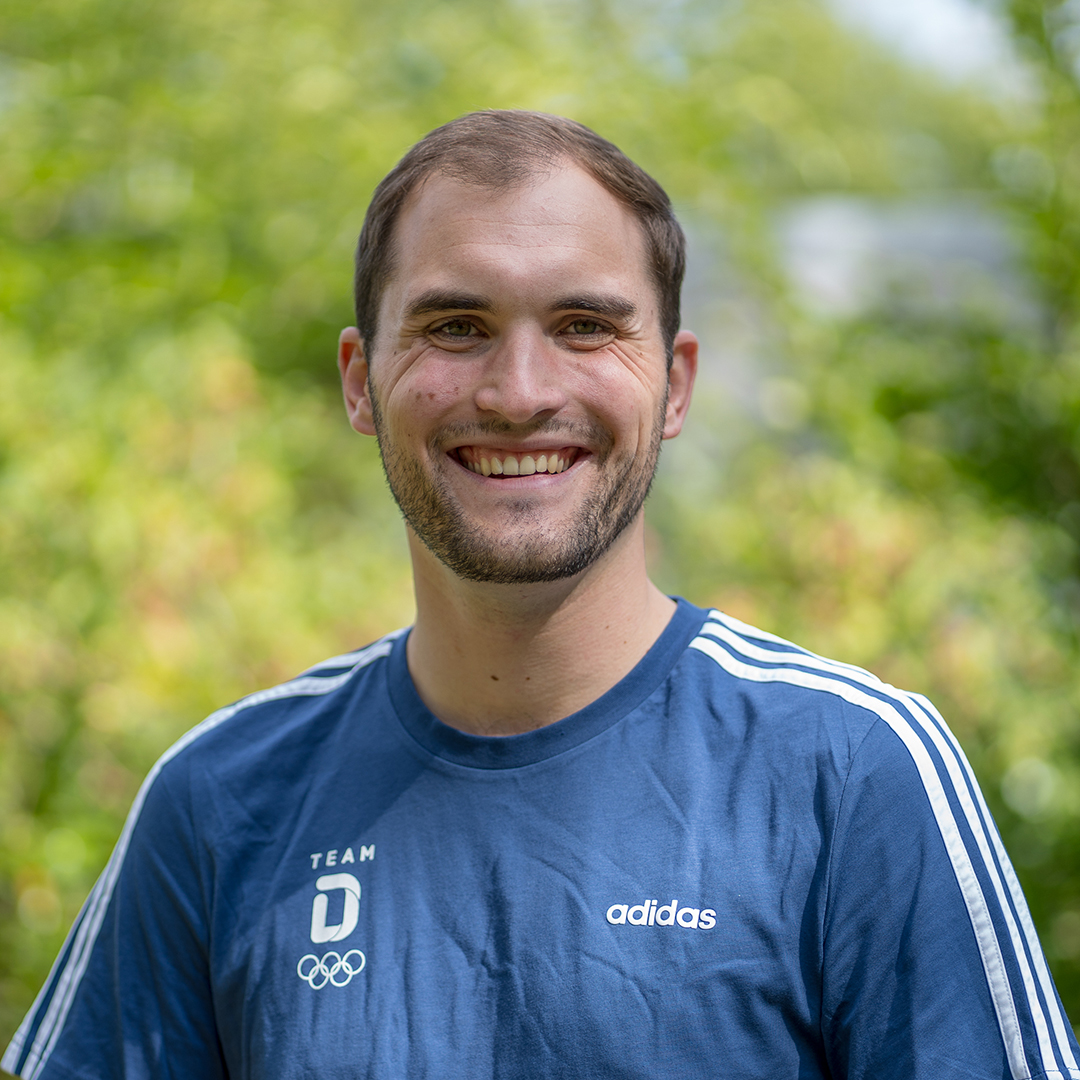 Porträt Luca Wernert mit kurzen Haaren trägt ein blaues T-Shirt mit weißen Streifen und dem Logo von Adidas. Er steht im Freien vor einer grünen, unscharfen Hintergrundlandschaft.