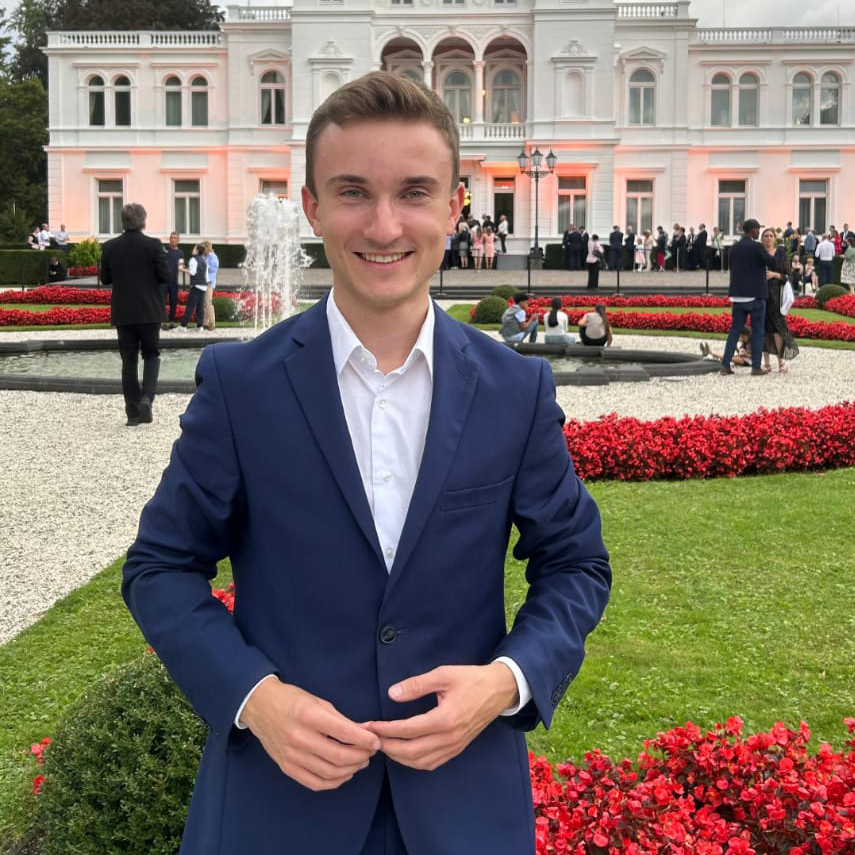 Porträt von Tom Faßhauer in einem blauen Anzug steht lächelnd vor einem eleganten Gebäude mit Garten und Blumenbeeten. Im Hintergrund sind weitere Personen zu sehen.