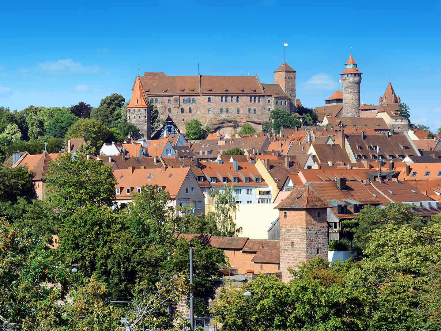 Blick auf eine historische Stadt mit roten Dächern und einer beeindruckenden Burg im Hintergrund. Die Burg hat mehrere Türme und ist von Bäumen umgeben. Der Himmel ist klar und blau.