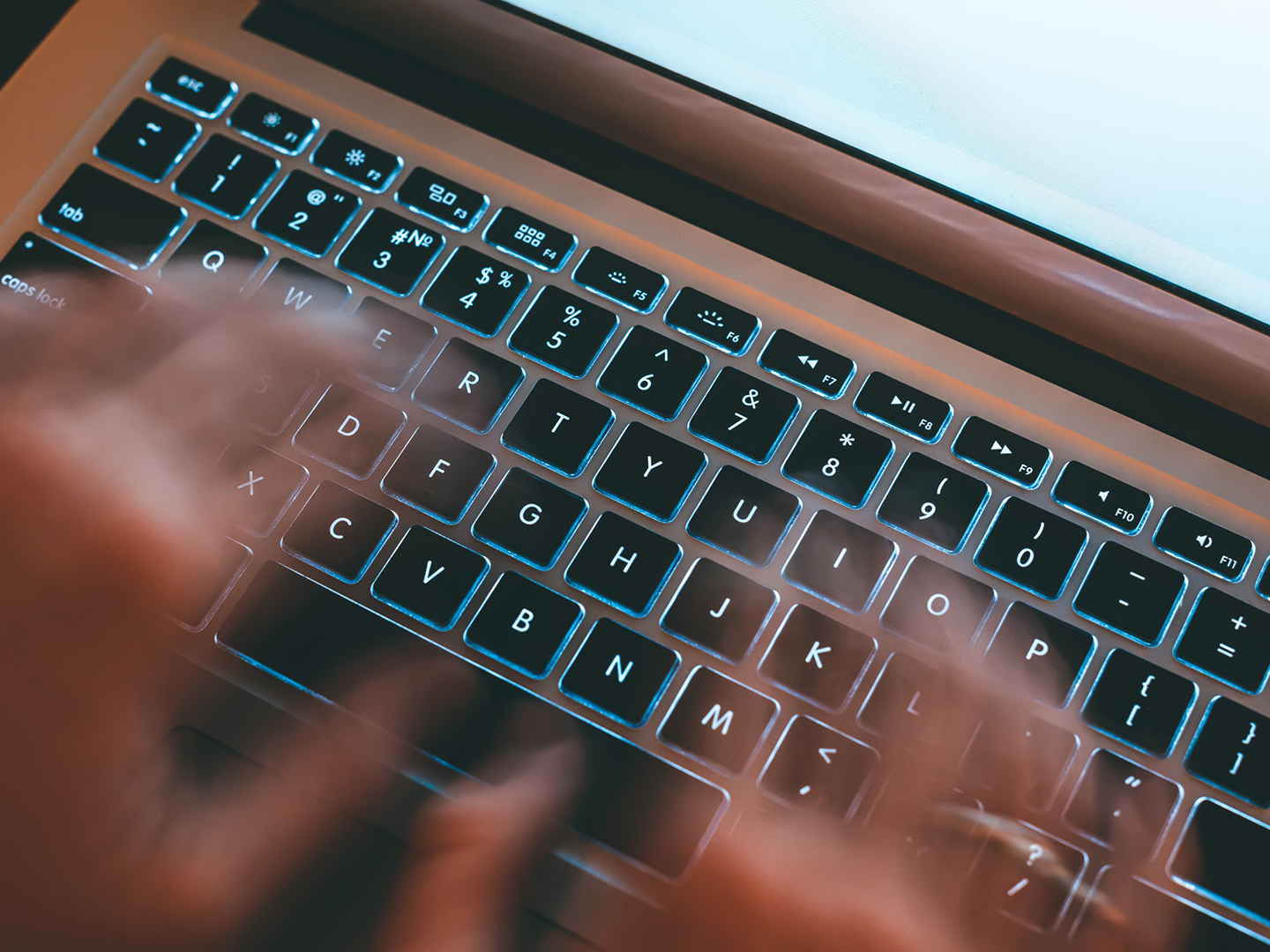 Eine verschwommene Ansicht von Händen, die schnell über eine beleuchtete Laptop-Tastatur tippen. Die Tasten sind deutlich sichtbar, während der Hintergrund unscharf ist.