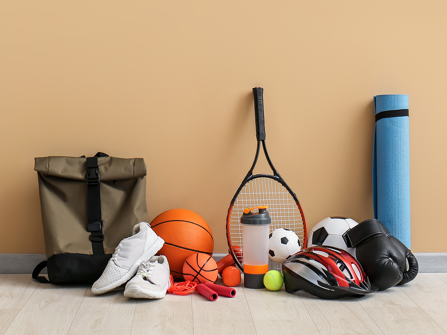 Eine Sporttasche, ein Paar Turnschuhe, ein Basketball, ein Tennisracket, eine Wasserflasche, ein Fußball, ein Tennisball, ein Paar Boxhandschuhe, ein Fahrradhelm und eine Yogamatte sind vor einer beige Wand angeordnet.