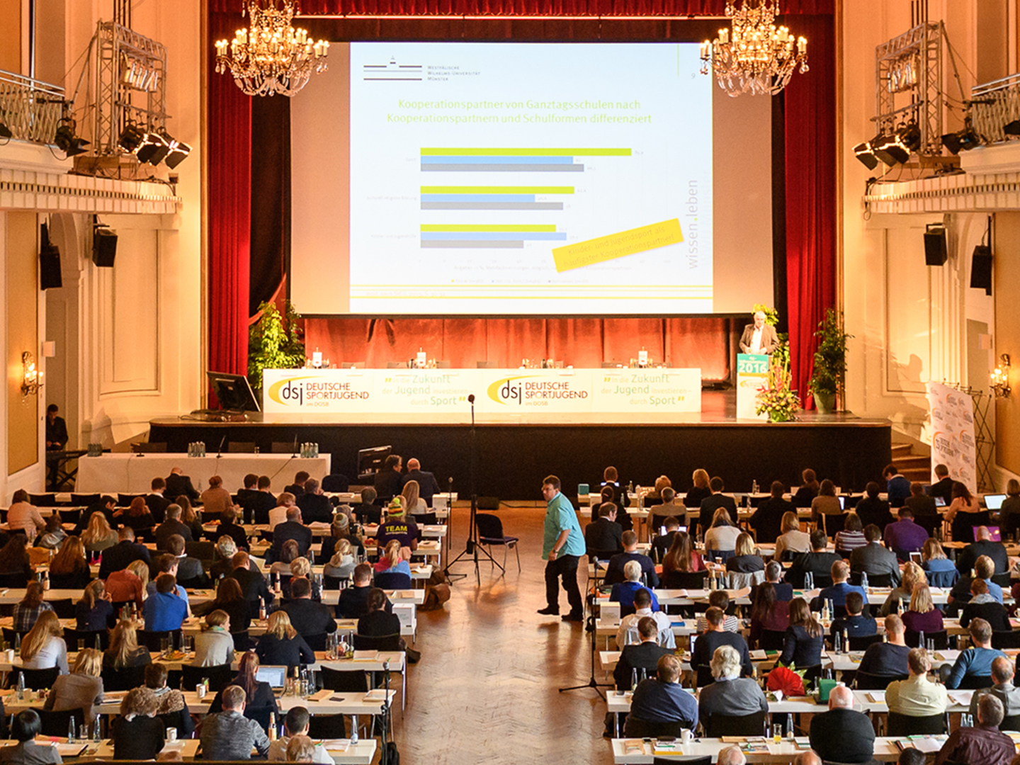 Eine große Konferenz mit vielen Teilnehmern in einem Saal. Auf der Bühne ist ein Präsentationsbildschirm mit Diagrammen zu sehen. Die Zuschauer sitzen an Tischen und schauen nach vorne. Der Raum ist festlich dekoriert mit Kronleuchtern und rotem Vorhang.