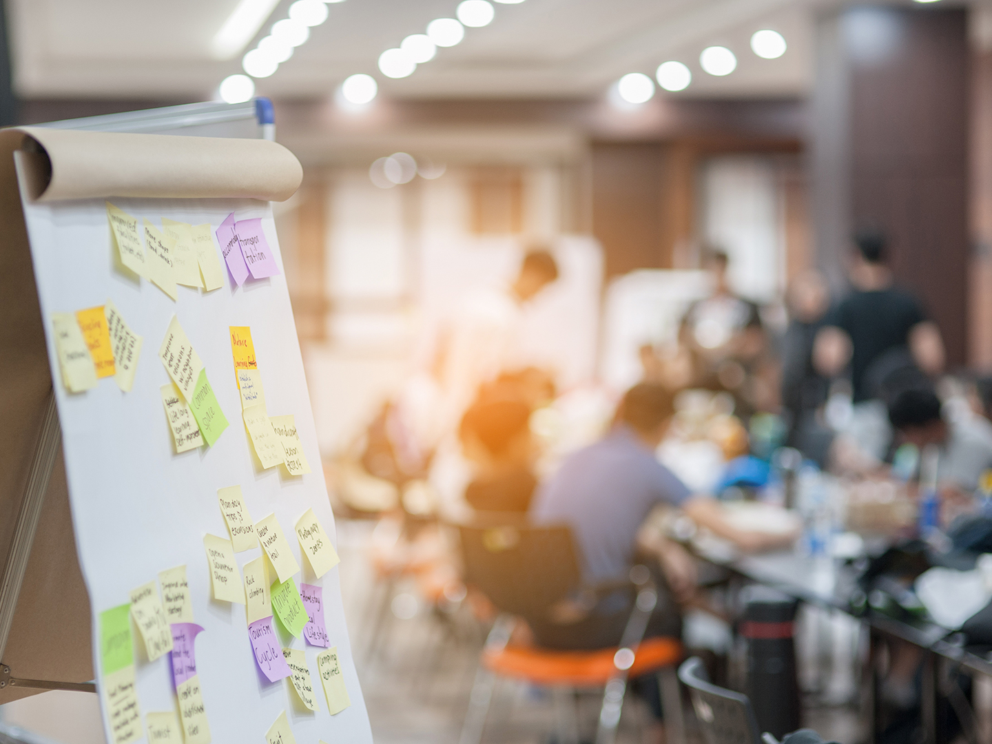 Ein Flipchart mit bunten Haftnotizen steht im Vordergrund. Im Hintergrund sind Menschen in einem Besprechungsraum zu sehen, die an Tischen sitzen und miteinander interagieren. Die Atmosphäre wirkt kreativ und kollaborativ.