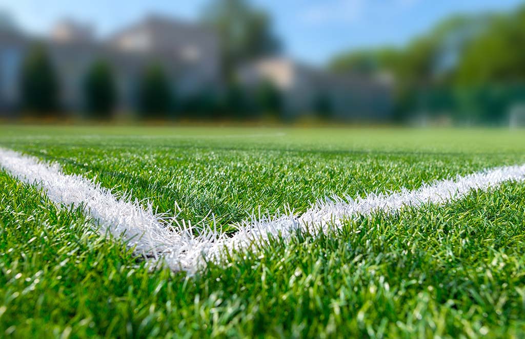 Nahaufnahme eines Fußballfeldes mit grünem Kunstrasen und einer weißen Linienmarkierung in der Ecke. Der Hintergrund ist unscharf und zeigt Bäume und Gebäude.