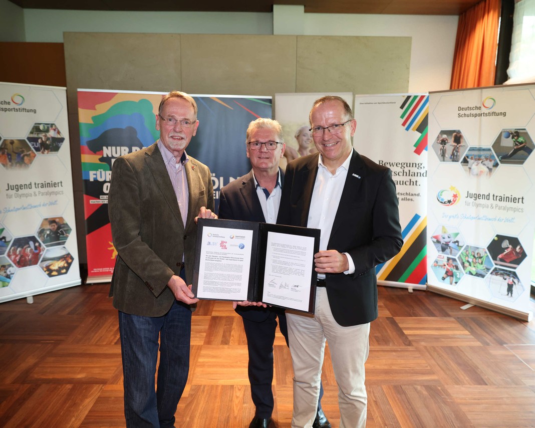 Drei Männer stehen in einem Raum und halten ein Dokument in den Händen. Im Hintergrund sind Banner mit Informationen über die Deutsche Schulsportstiftung und das Projekt "Jugend trainiert". Die Männer wirken erfreut und engagiert.