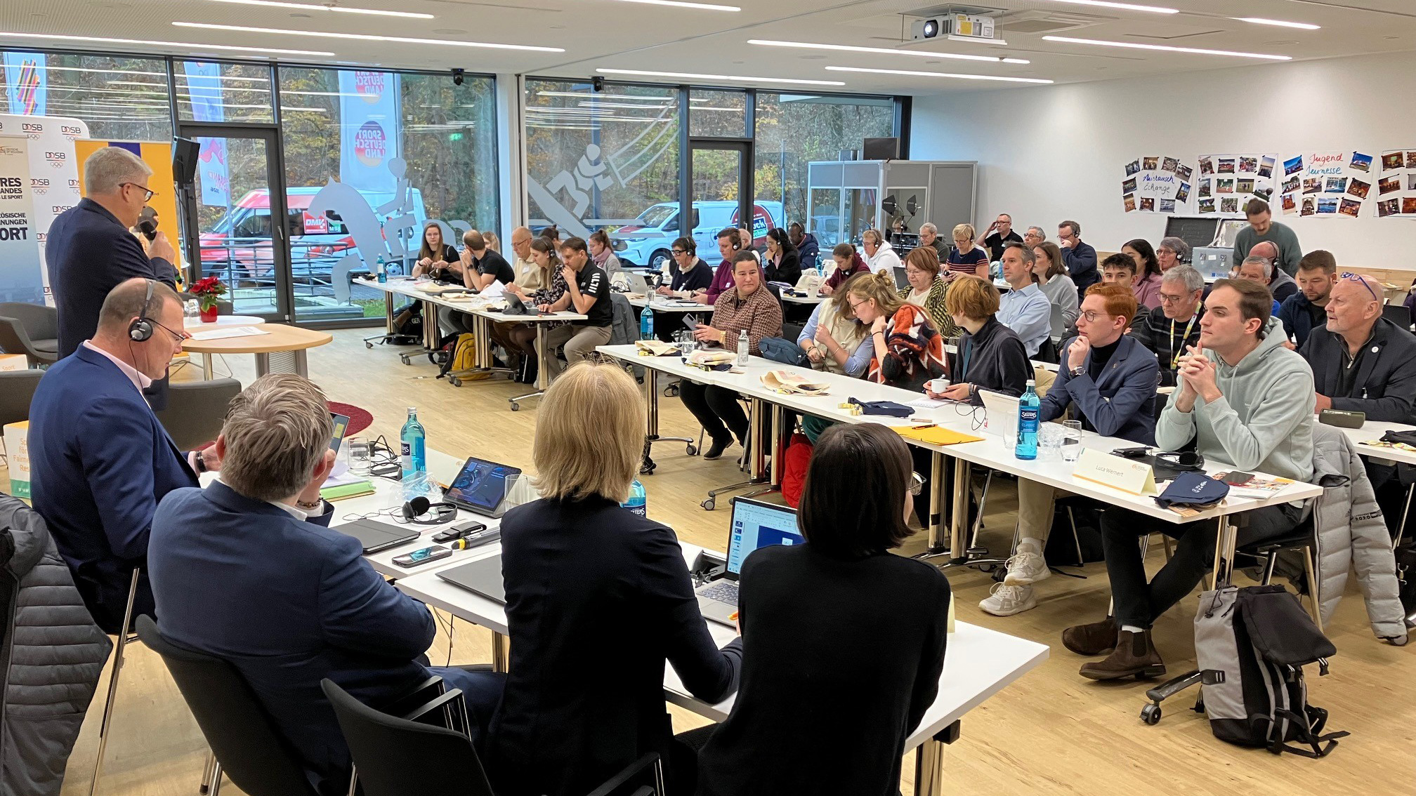 In einem hellen, modernen Seminarraum sitzen zahlreiche Personen an langen Tischen, die in Reihen angeordnet sind. Im Vordergrund sind vier Personen von hinten zu sehen, die an einem Tisch mit Laptops und Wasserflaschen sitzen. Im Hintergrund steht ein Mann an einem Rednerpult und spricht zu den Anwesenden. Große Fenster an der linken Seite lassen Tageslicht herein und bieten Blick auf geparkte Autos. An der rechten Wand sind zahlreiche Fotos oder kleine Plakate angeordnet. Die Atmosphäre wirkt formell und konzentriert, passend zu einer Tagung.