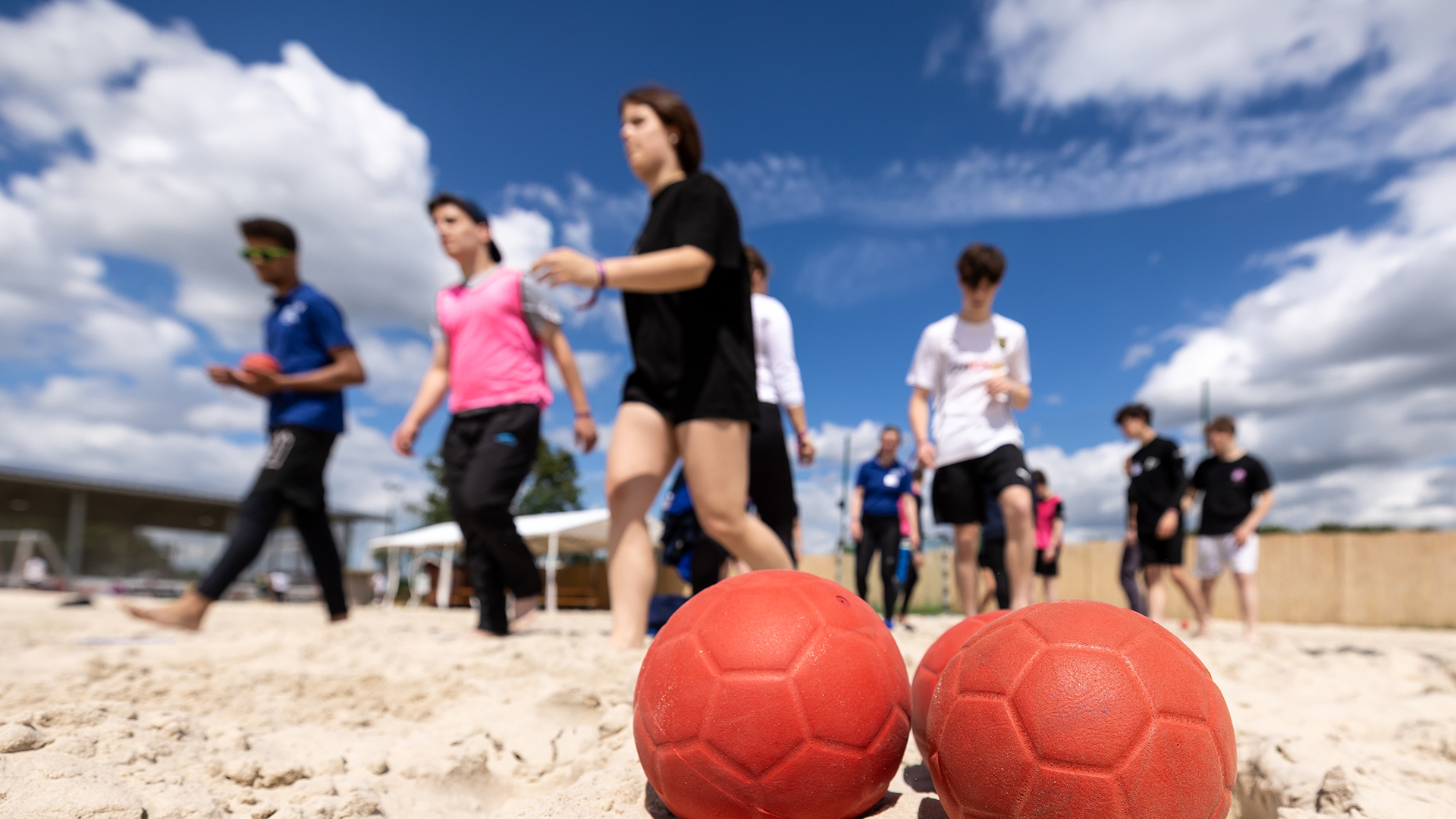 Mehrere Jugendliche laufen auf einem Sandplatz. Im Vordergrund liegen zwei rote Bälle auf dem Sand. Die Jugendlichen tragen sportliche Kleidung in verschiedenen Farben, darunter ein schwarzes T-Shirt und Shorts, ein pinkes T-Shirt sowie ein weißes T-Shirt. Der Himmel ist blau mit einigen Wolken, im Hintergrund ist eine Brücke zu sehen.