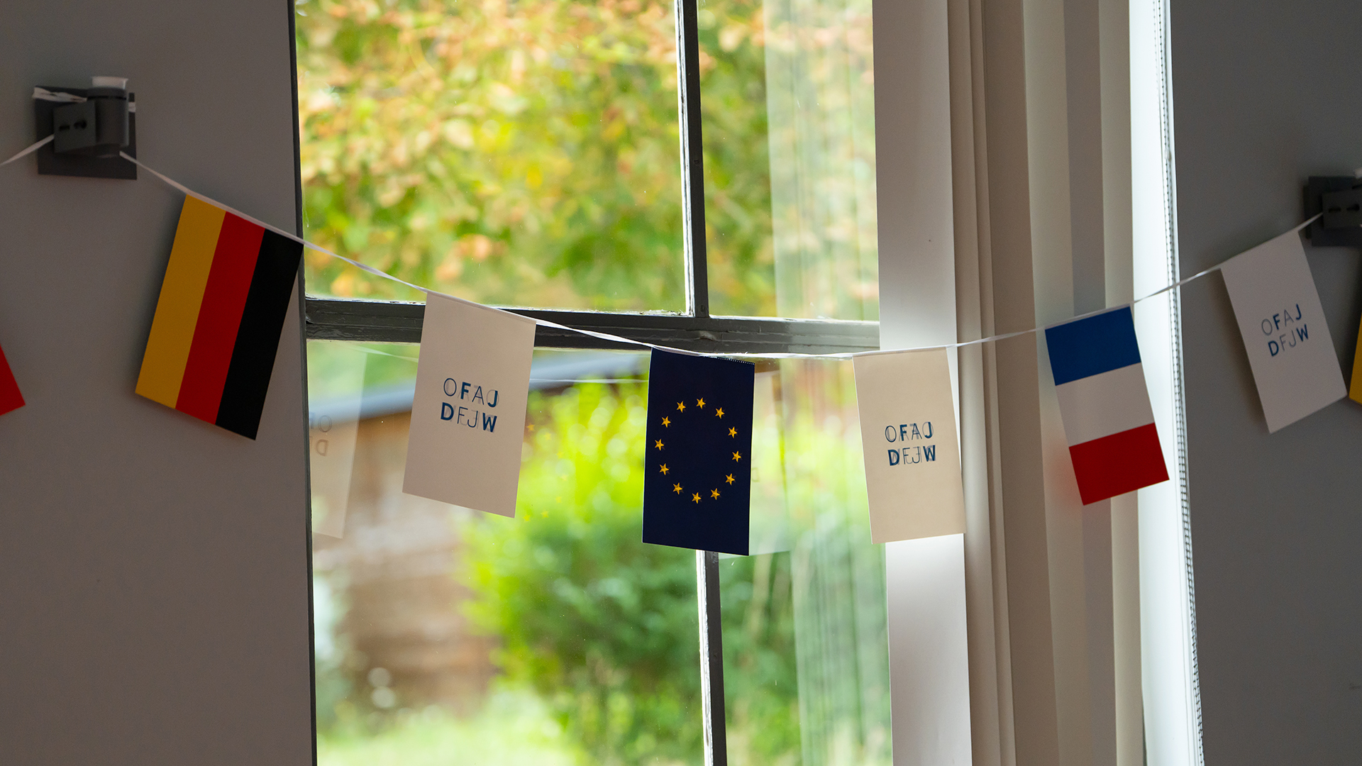 Girlande mit Deutschland-, EU- und Frankreich-Flaggen sowie zwei weiße Flaggen mit der Aufschrift 'OFAJ DFJW' vor einem Fenster