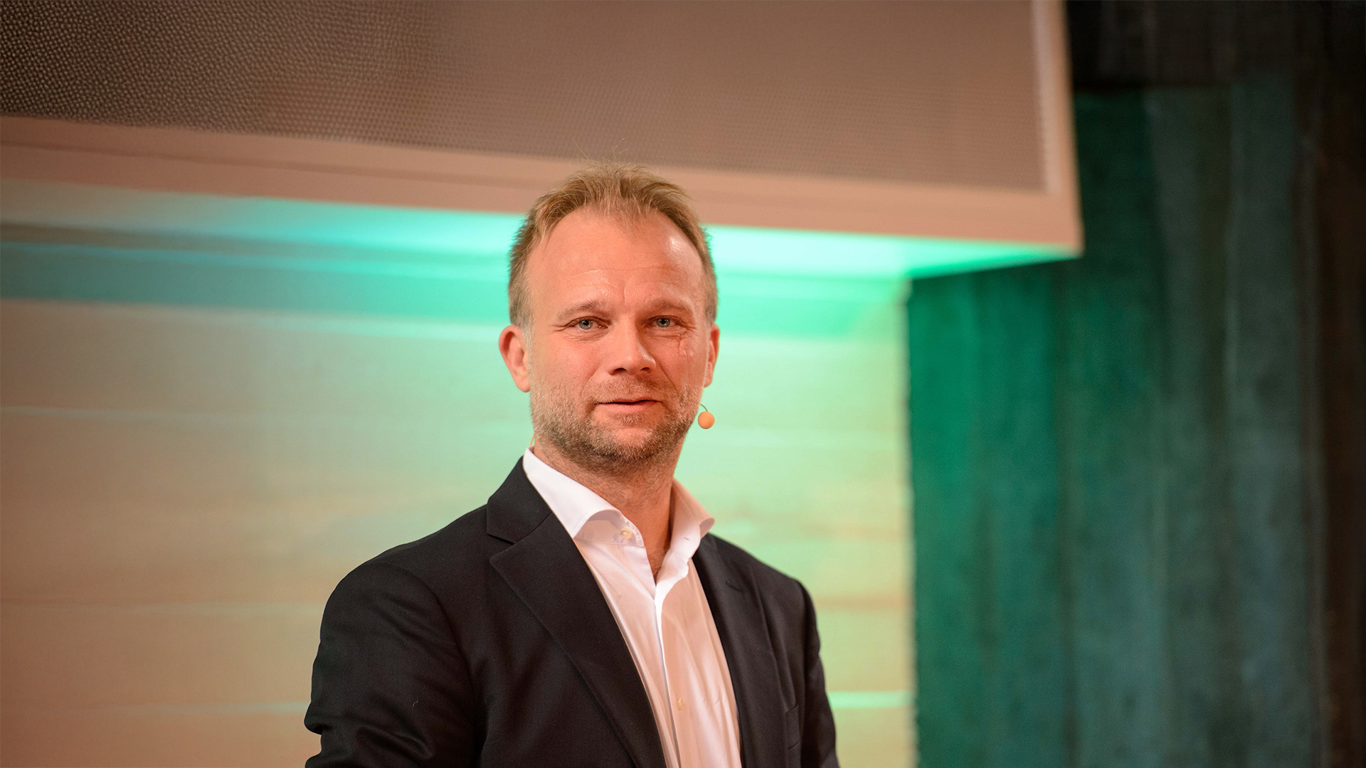 Benny Folkmann steht auf einer Bühne vor einem grün beleuchteten Hintergrund und trägt ein dunkles Sakko mit hellem Hemd.