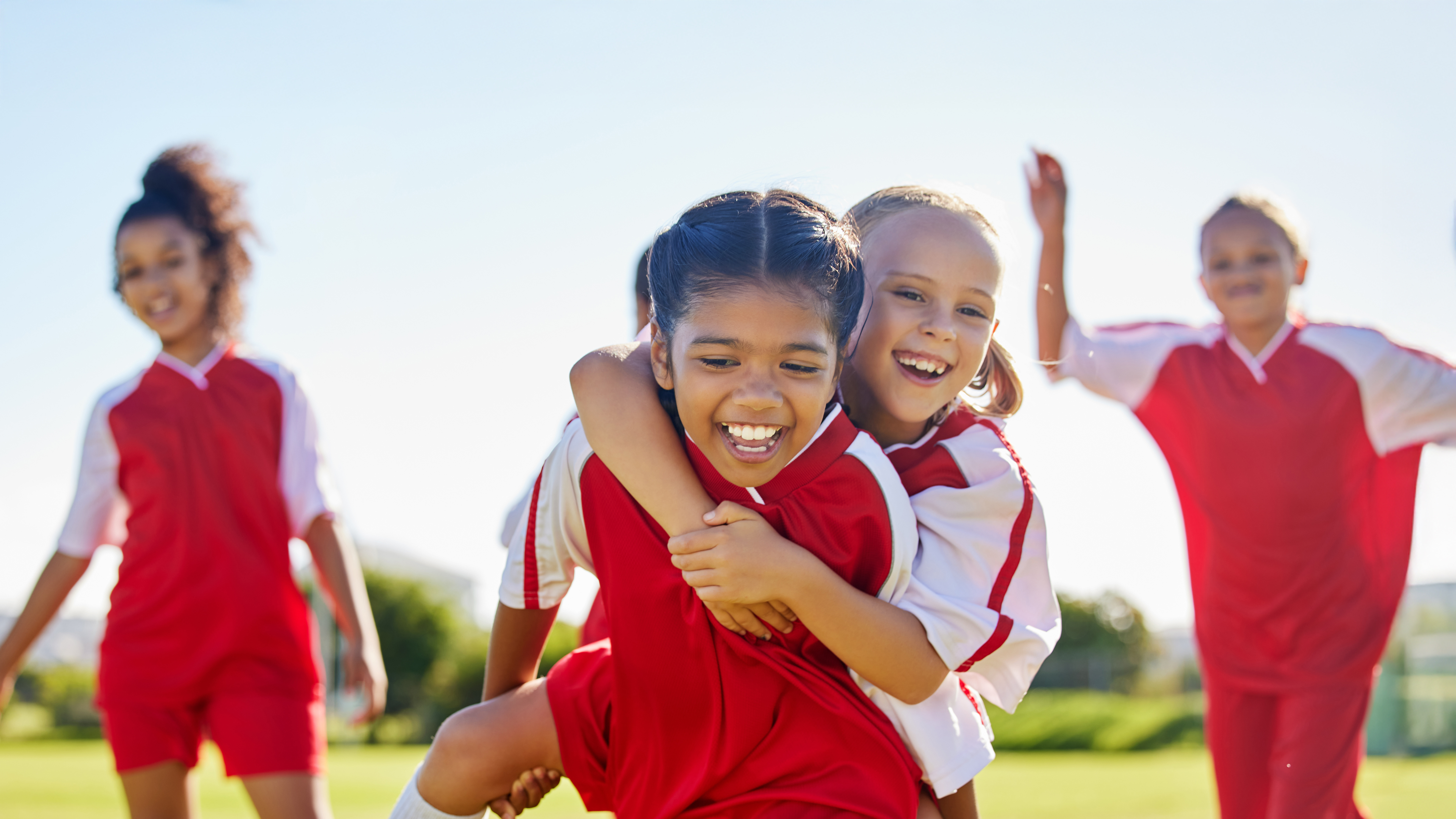 Zwei Kinder in rot-weißen Sporttrikots umarmen sich auf einem grünen Sportplatz, während zwei weitere Kinder im Hintergrund stehen. Die Kinder tragen weiße Sportsocken und Sportschuhe. Die Szene spielt bei Tageslicht im Freien.