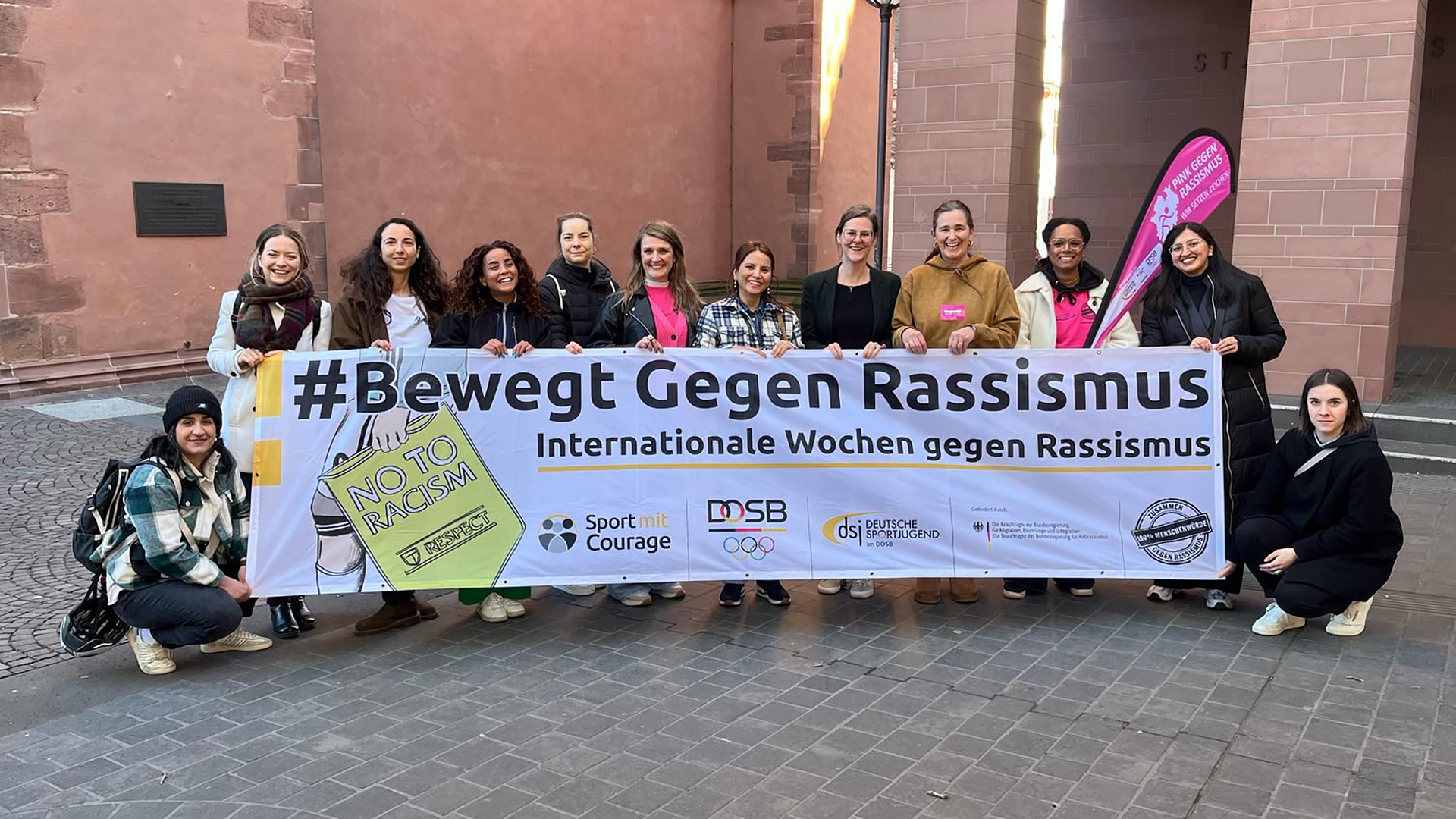 Eine Gruppe von jungen Menschen steht vor einem Gebäude und hält gemeinsam ein großes Banner mit der Aufschrift „#BewegtGegenRassismus – Internationale Wochen gegen Rassismus“, ergänzt durch Logos von Sport‑ und Jugendorganisationen.