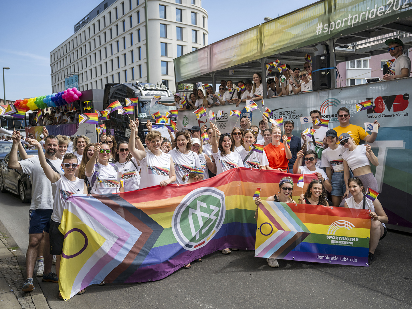 Gruppe von Teilnehmer*innen beim CSD Frankfurt mit Regenbogenflaggen und einem Banner. Sie tragen T-Shirts mit der Aufschrift „Loud and Proud im Sport“ und posieren vor einem bunt geschmückten Festwagen der Parade.