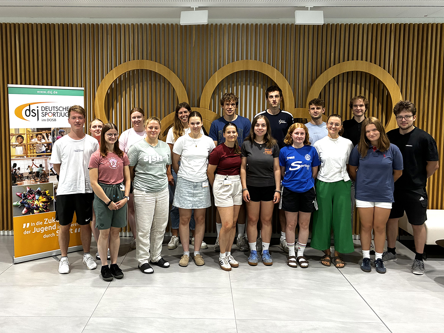 Eine Gruppe von 18 jungen Erwachsenen steht in einem modernen Raum. Sie lächeln und posieren gemeinsam. Im Hintergrund ist ein Banner der Deutschen Sportjugend zu sehen. Die Personen tragen verschiedene Freizeitkleidung und stehen in mehreren Reihen.