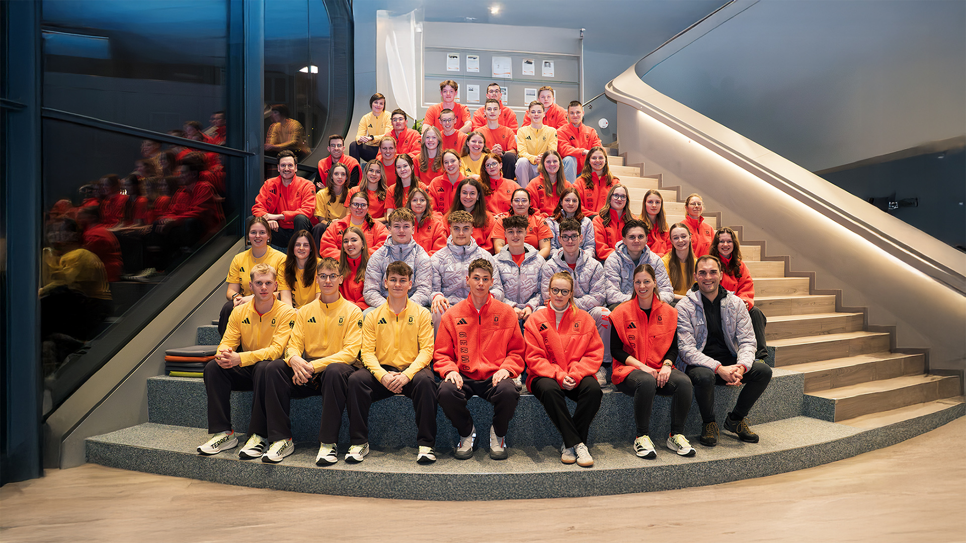 Gruppenfoto von 40 Jugendlichen zum Start des Deutschen Olympischen Jugendlagers (DOJL) Milano Cortina 2026; Teilnehmende in farbigen Teamjacken sitzen auf einer modernen Treppe im Veranstaltungsgebäude beim Auftakt unter dem Motto „It’s Our Vibe“.