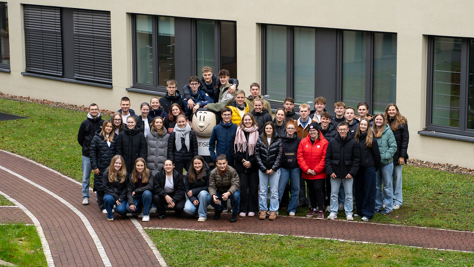 Gruppenfoto der Teilnehmenden und des Leitungsteams des Deutschen Olympischen Jugendlagers Milano Cortina 2026 beim Vortreffen: Eine große Gruppe von Menschen steht dicht beieinander vor einem modernen Gebäude mit grauen Wänden und mehreren Fenstern mit geschlossenen Jalousien. Die Personen tragen überwiegend dunkle, winterliche Jacken, einige auch helle oder rote. Sie befinden sich auf einem gepflasterten Weg und einem angrenzenden Rasenstück. 