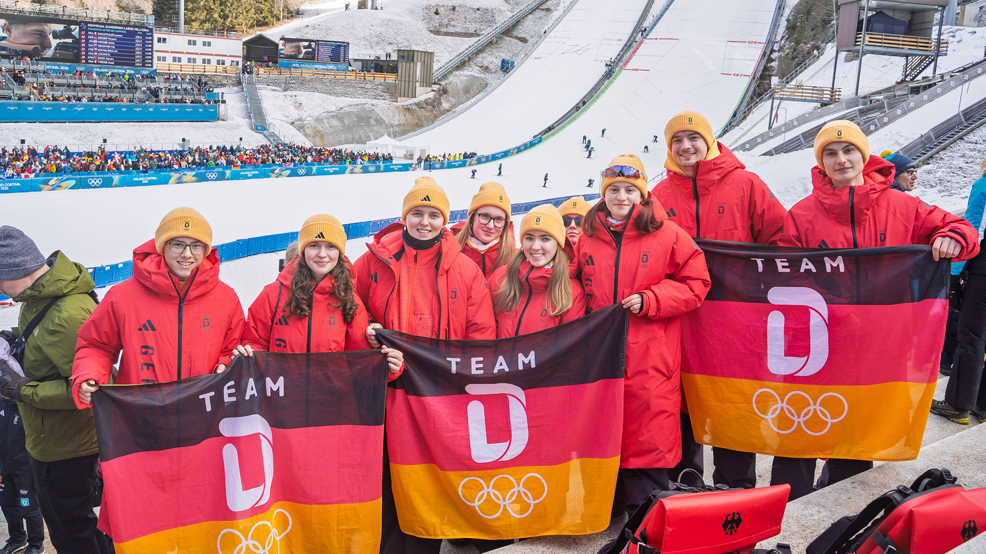Gruppe junger Menschen aus dem Deutschen Olympischen Jugendlager Milano Cortina 2026 in roten Teamjacken hält Deutschland‑Fahnen mit olympischem Emblem in einem winterlichen Sportstadion; im Hintergrund sind eine Skisprungschanze und Zuschauerränge zu sehen.