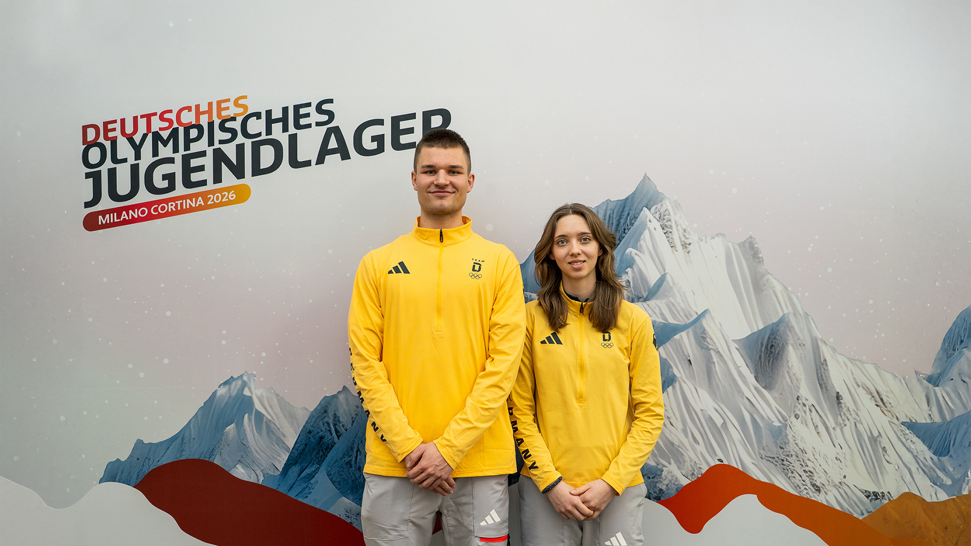 Zwei Personen in gelben Sportjacken des Deutschen Olympischen Jugendlagers stehen vor einer Wand mit dem Logo „Deutsches Olympisches Jugendlager Milano Cortina 2026“ und einer grafischen Berglandschaft.