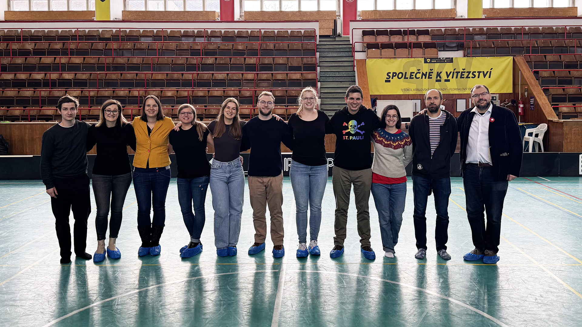 Gruppenfoto beim deutsch-tschechischen Fachkräfteaustausch in Prag mit dem tschechischen Breitensportverband Sokol. Elf Personen stehen in einer Sporthalle nebeneinander in einer Reihe. Sie tragen unterschiedliche Kleidung, darunter ein gelber Pullover, ein schwarzes T-Shirt mit buntem Aufdruck, ein blau-weiß-roter Pullover und ein weißes Hemd mit schwarzem Sakko. Im Hintergrund sind leere Tribünen mit roten und braunen Sitzplätzen sowie ein gelbes Banner mit schwarzer Schrift in tschechischer Sprache zu sehen. Der Austausch wird im Rahmen eines Kooperationsprojekts mit TANDEM, dem Koordinierungszentrum für den deutsch-tschechischen Jugendaustausch, durchgeführt.