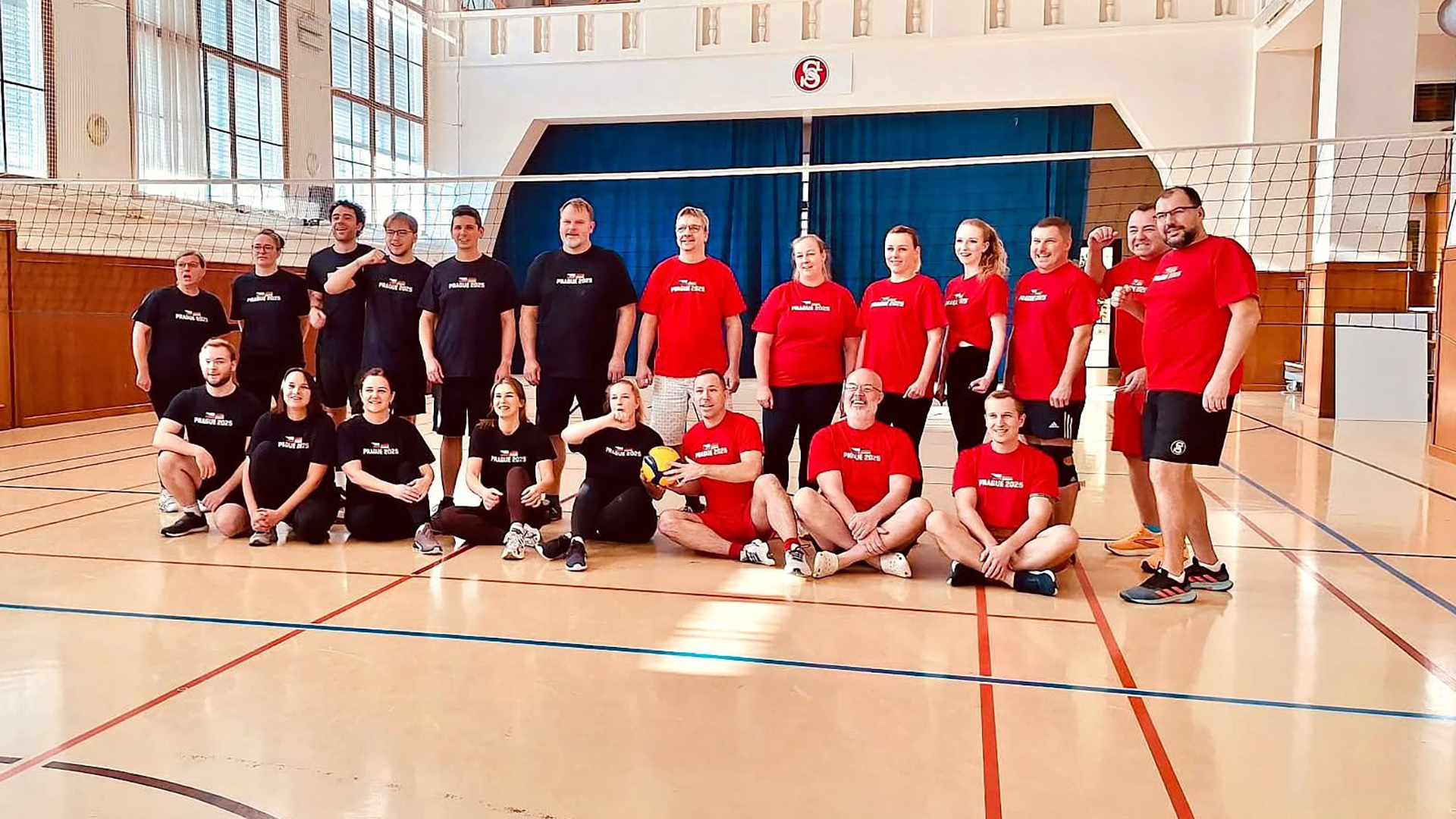 Gruppenfoto von Teilnehmenden des deutsch‑tschechischen Fachkräfteaustauschs in einer Sporthalle in Nürnberg: Fachkräfte stehen und sitzen gemeinsam auf dem Hallenboden, einige halten einen Ball, und tragen überwiegend einheitliche T‑Shirts – Ausdruck von Begegnung, Austausch und gemeinsamer sportlicher Praxis.