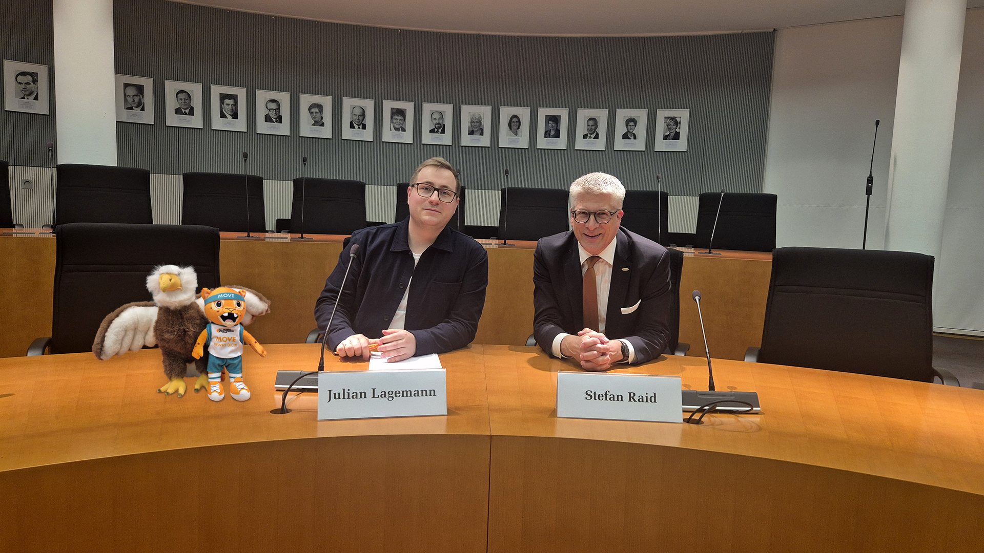 Deutsche Sportjugend als Sachverständige bei der Kinderkommission: Links die Maskottchen der Kinderkommission und Movelino von der dsj, daneben Julian Lagemann und Stefan Raid an einem Sitzungstisch.