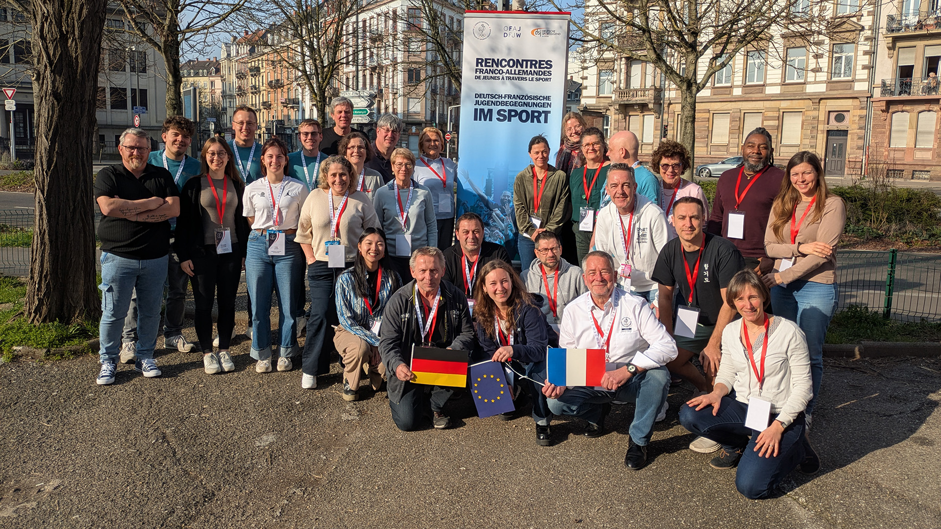 Gruppenfoto junger Erwachsener bei einer deutsch-französischen Infotagung im Sport: Die Teilnehmenden stehen und knien im Freien vor einem Banner „Rencontres franco-allemandes de la jeunesse – Jugendbegegnungen im Sport“ und halten unter anderem deutsche, französische und EU-Flaggen; im Hintergrund sind Stadtbäume und Gebäude zu sehen.