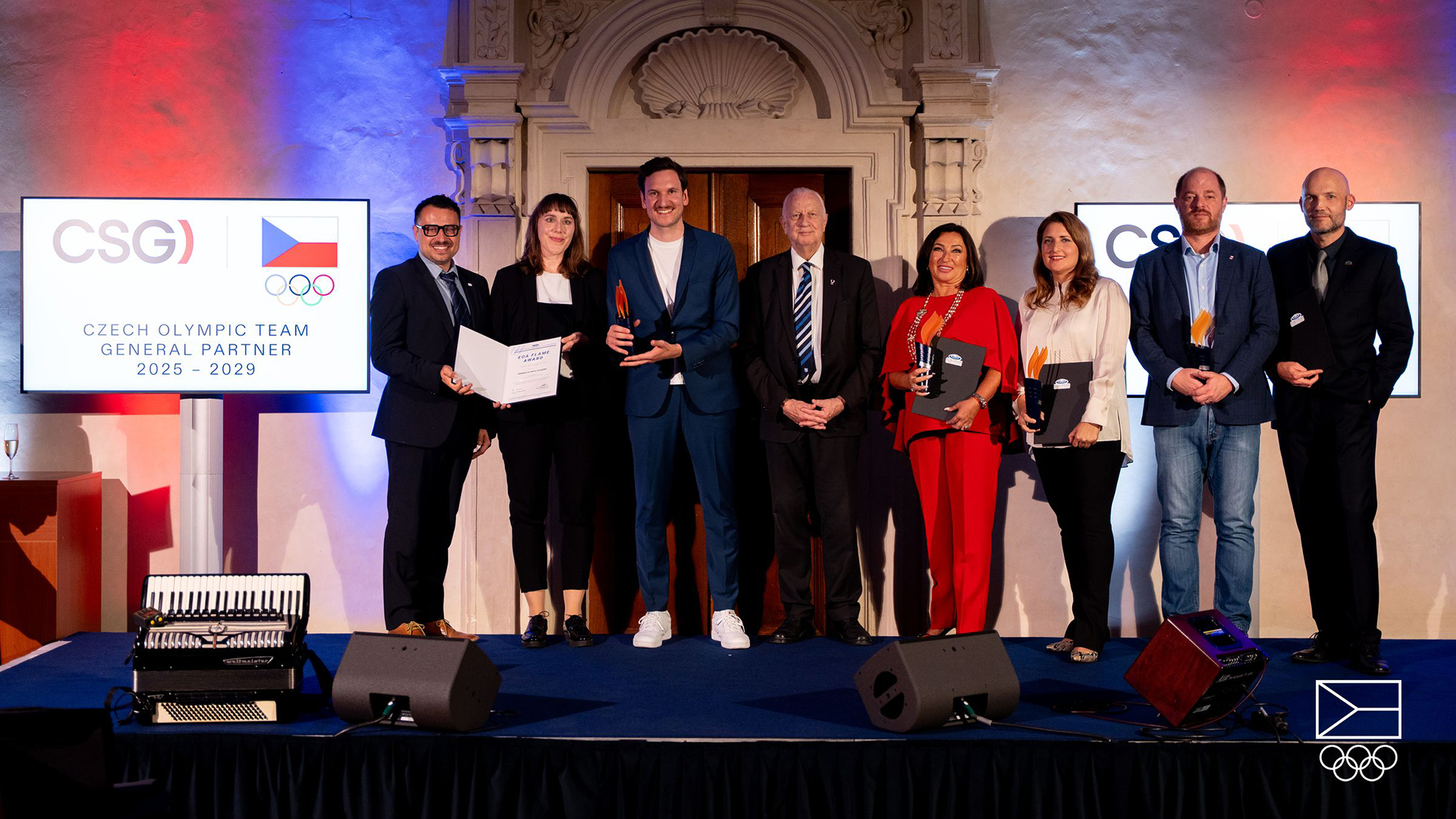 Personengruppe auf Bühne bei Flame Award für DFOJL Paris 2024, einige halten Urkunden und Trophäen, zwei Bildschirme mit Logo und Text 'Czech Olympic Team General Partner 2025 - 2029' im Hintergrund.