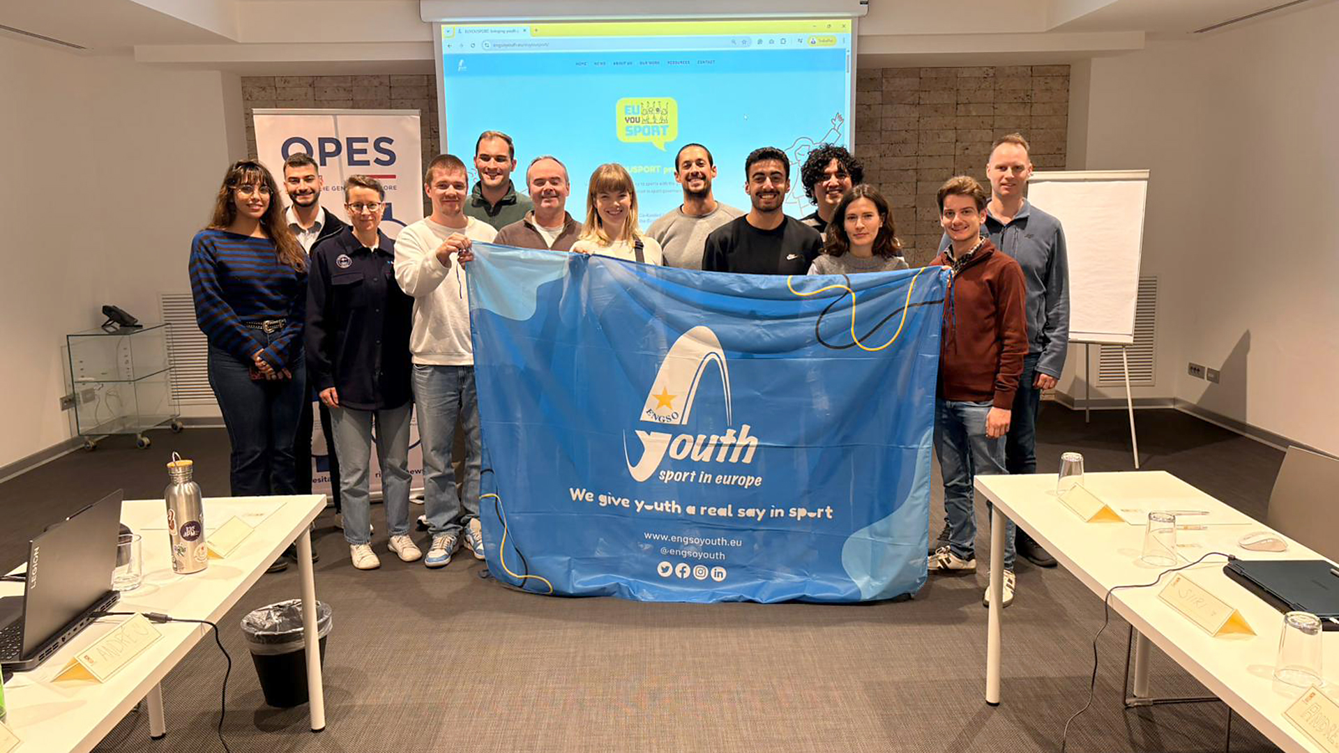 Gruppenfoto von Teilnehmenden des EU-Projekt EUYOUSPORT in einem Seminarraum, die gemeinsam ein blaues Banner mit dem Logo und dem Schriftzug „Youth Sport in Europe“ präsentieren; im Hintergrund sind eine Präsentationsfläche und Workshop-Materialien zu sehen.