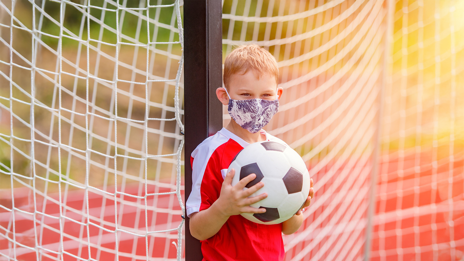 Ein Junge mit einer Corona-Maske im Gesicht in einem roten Fußballtrikot hält einen schwarz-weißen Fußball vor einem Fußballtor. Das Tor ist mit einem Netz ausgestattet.