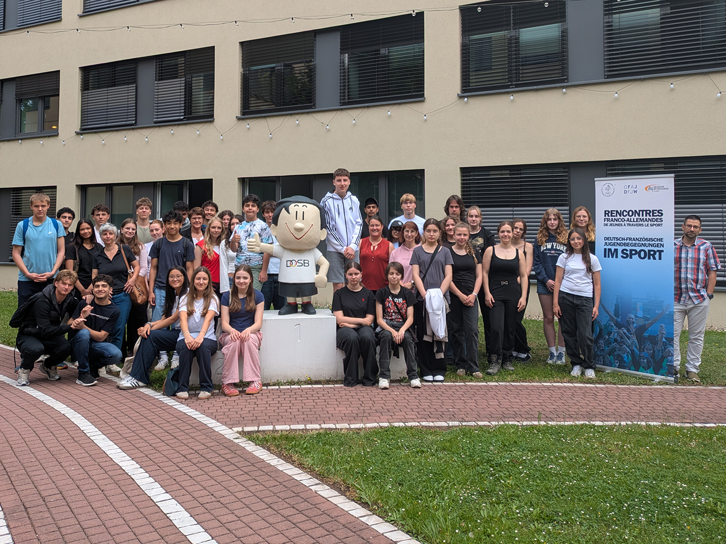Eine Gruppe von etwa 50 Personen steht vor einem Gebäude, umgeben von Rasen und einem Weg. In der Mitte steht ein Maskottchen. Rechts ist ein Banner mit dem Text "Deutsch-Französische Jugendbegegnungen im Sport" zu sehen. Die Teilnehmer sind in verschiedenen Kleidungsstilen gekleidet.