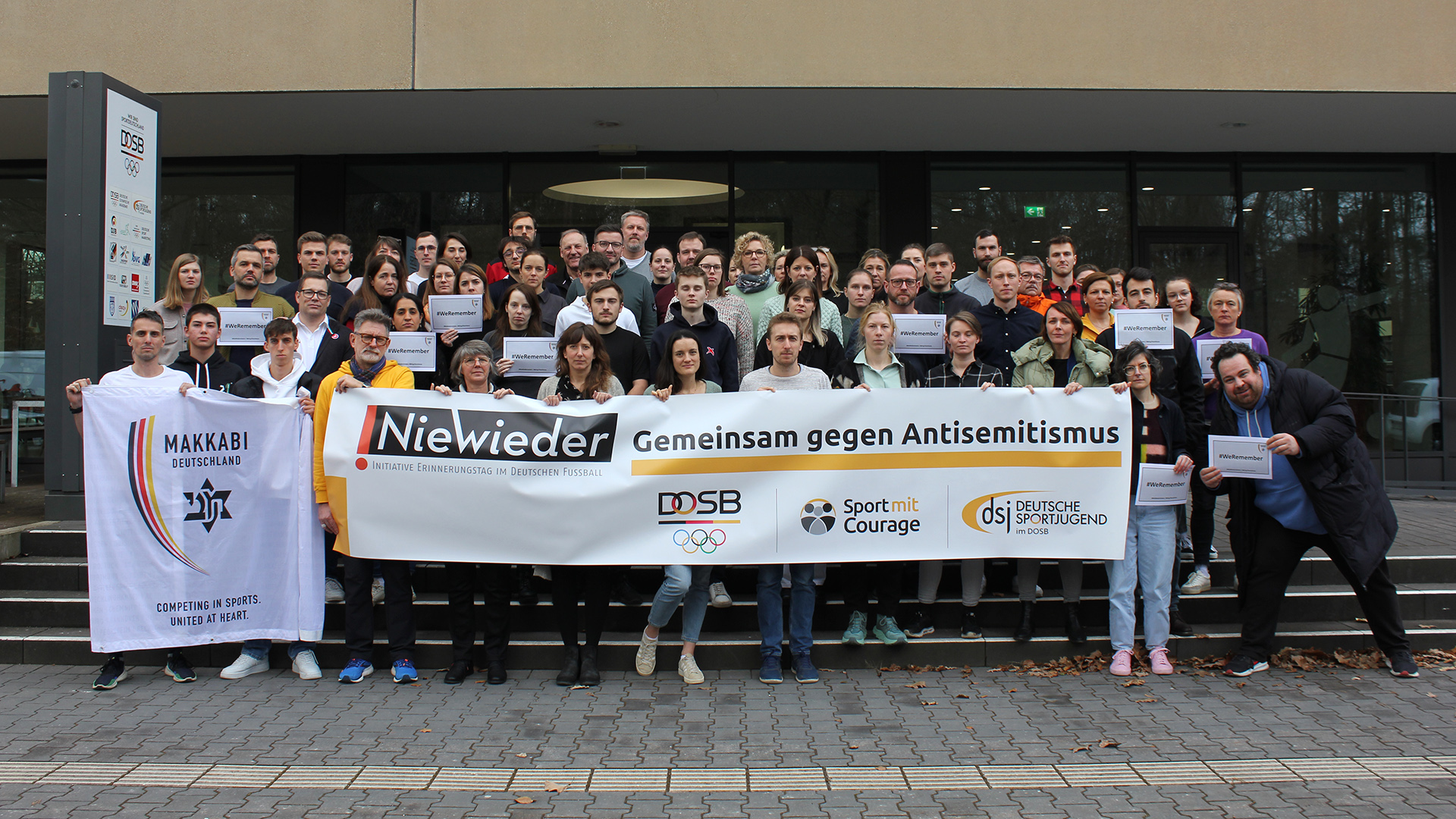 Gruppenfoto vor einem Gebäude mit einem großen Banner der Initiative ‚!NieWieder – Gemeinsam gegen Antisemitismus‘, unterstützt von DOSB, Deutsche Sportjugend, Sport mit Courage und Makkabi Deutschland. Symbolbild für den Fach- und Begegnungstag ‚Antisemitismus im Fußball‘ im Deutschen Fußballmuseum und das Engagement des Sports gegen Antisemitismus.