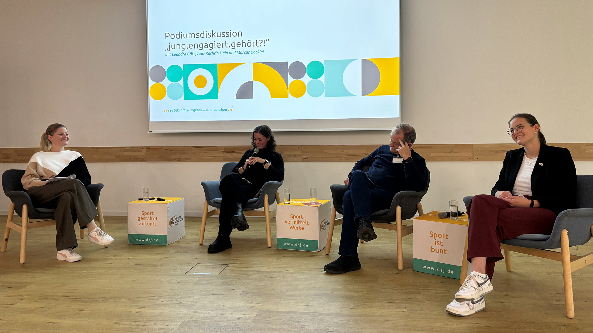 Podiumsdiskussion mit vier Personen auf Stühlen, drei Würfel mit Aufschriften 'Sport gestaltet Zukunft', 'Sport verbindet Welten' und 'Sport ist bunt', im Hintergrund Projektion mit Titel 'Podiumsdiskussion „jung.engagiert.gehört?“