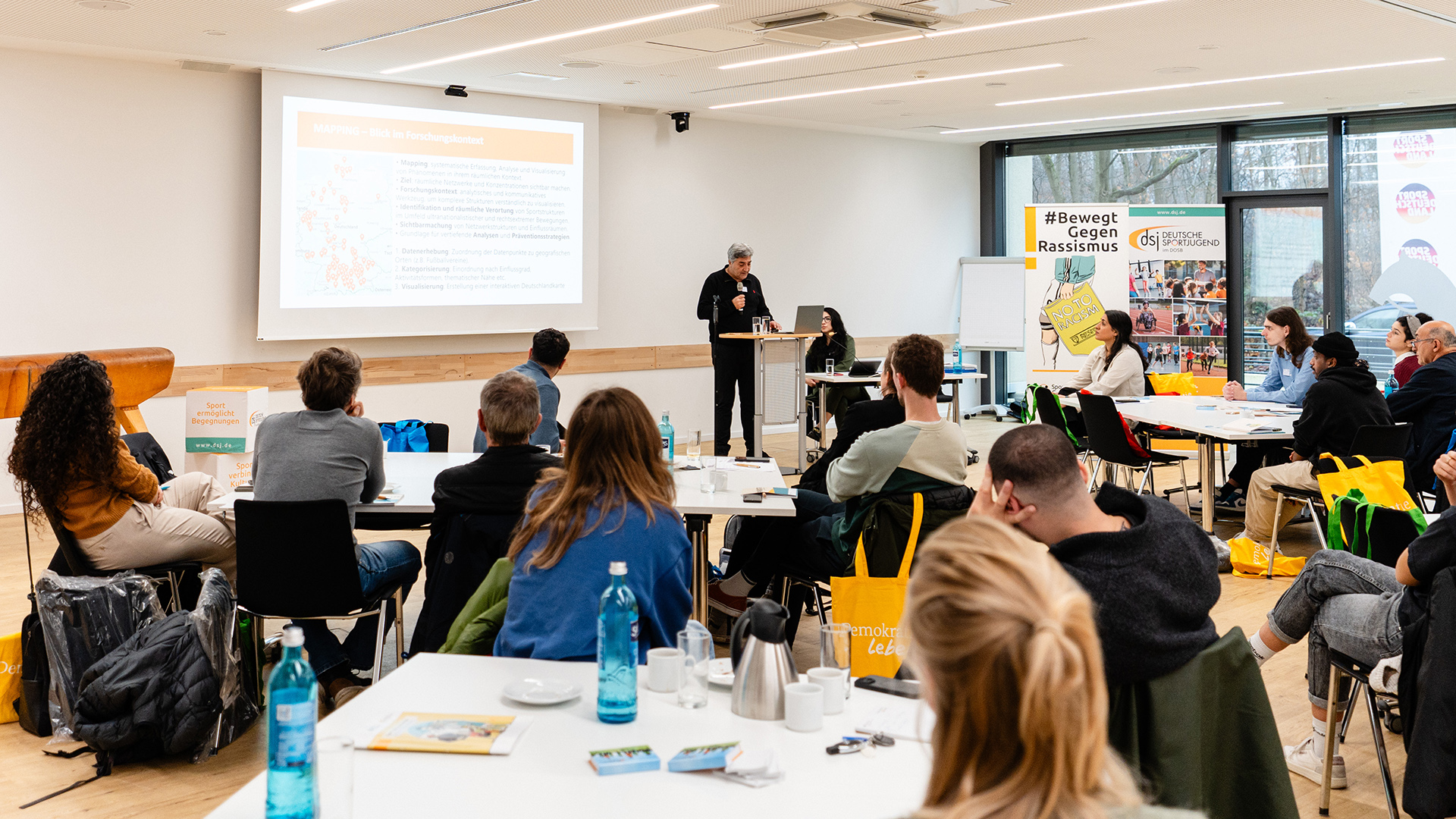 In einem hellen, modernen Seminarraum steht eine Person an einem Rednerpult und hält einen Vortrag. Vor der Person sitzen mehrere Zuhörende an Tischen, die mit Wasserflaschen, Kaffeekannen und Unterlagen ausgestattet sind. Im Hintergrund ist eine große Leinwand mit einer Präsentation zu sehen. Rechts im Raum befinden sich zwei große Plakate, eines davon trägt die Aufschrift '#Bewegt Gegen Rassismus'. Die Zuhörenden sind in lockerer Sitzordnung verteilt und hören aufmerksam zu.