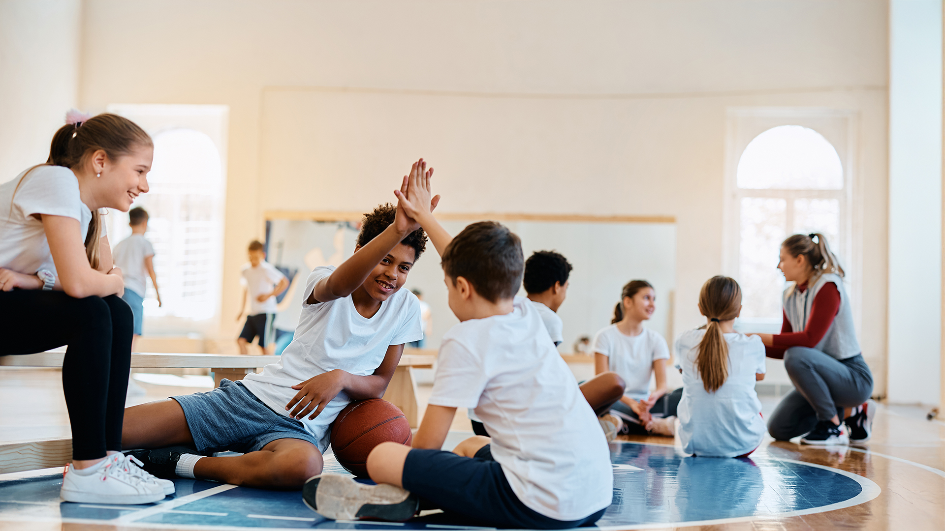 In einem hellen Raum mit großen Fenstern sitzen mehrere Kinder in Sportkleidung auf einem Hallenboden, der mit einem blauen Kreis markiert ist. Zwei Kinder geben sich in der Bildmitte einen High-Five. Im Hintergrund sitzt eine erwachsene Person in sportlicher Kleidung auf dem Boden und spricht mit einer Gruppe von Kindern. Die Atmosphäre wirkt freundlich und aktiv.
