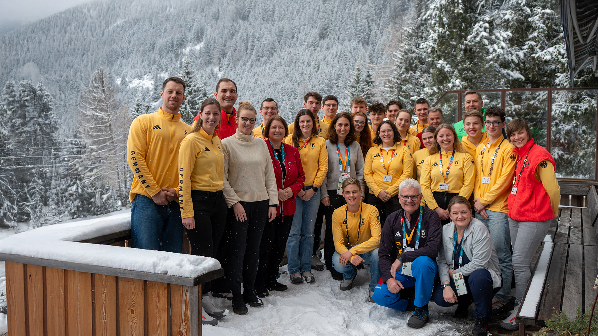 Gruppe junger Teilnehmender des Deutschen Olympischen Jugendlagers steht gemeinsam mit Mitgliedern des Ausschusses für Sport und Ehrenamt, darunter Aydan Özoğuz, auf einer verschneiten Terrasse vor einer winterlichen Berglandschaft; viele tragen gelbe Teamjacken und Akkreditierungen.