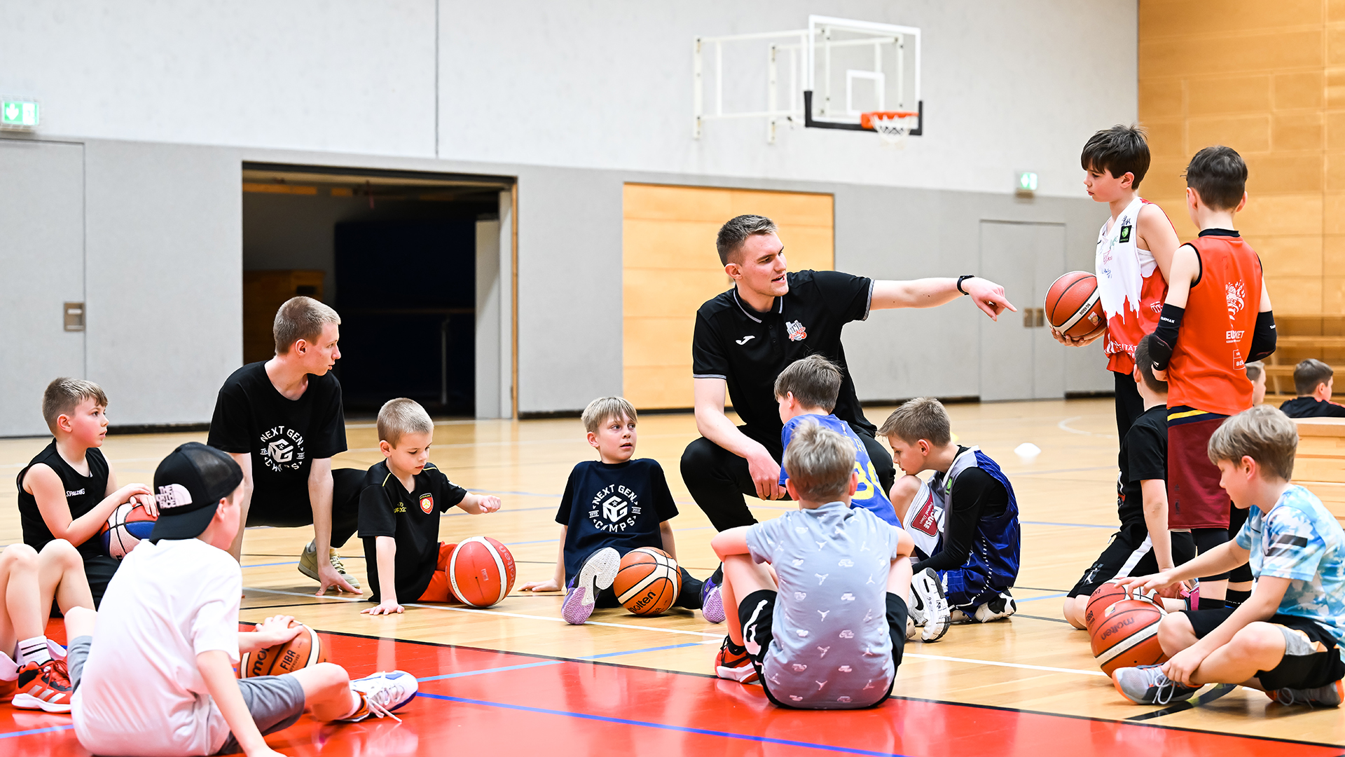In einer Sporthalle sitzt eine Gruppe von Kindern im Kreis auf dem Hallenboden, umgeben von Basketballen. Ein Erwachsener in schwarzer Sportkleidung sitzt mit ihnen und zeigt mit dem Arm nach rechts, während er spricht. Die Kinder tragen sportliche Kleidung in verschiedenen Farben, darunter T-Shirts und Shorts. Im Hintergrund ist ein Basketballkorb an der Wand zu sehen. Die Szene vermittelt eine Trainings- oder Unterrichtssituation im Basketball.