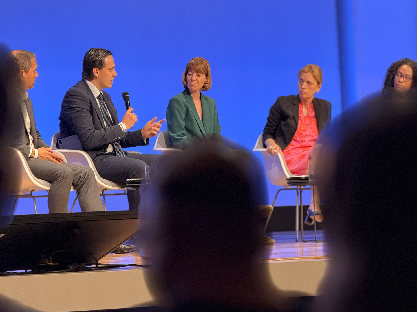 Eine Diskussionsrunde auf einer Bühne mit mehreren Personen. Ein Mann spricht ins Mikrofon, während die anderen Teilnehmer aufmerksam zuhören. Der Hintergrund ist blau beleuchtet, und die Zuschauer sind unscharf im Vordergrund zu sehen.
