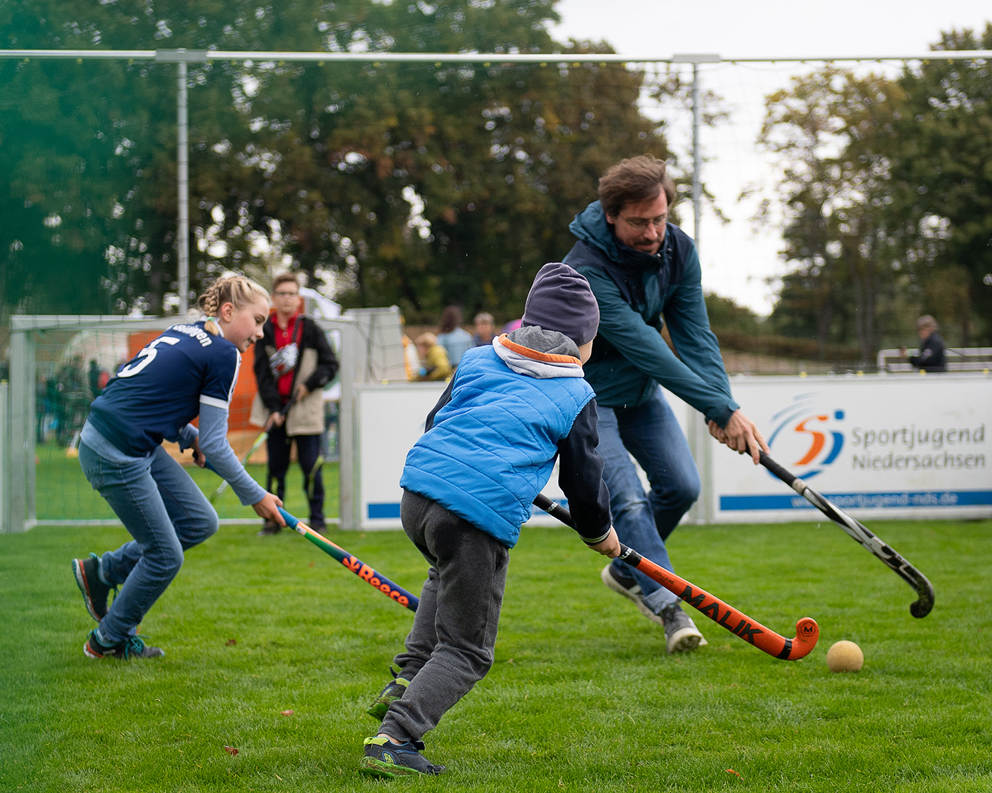 Drei Personen spielen Hockey auf einem Rasenplatz. Zwei Kinder, eines in blauer Jacke und eines in einem blauen T-Shirt, versuchen, den Ball zu erreichen, während ein Erwachsener mit einem Schläger in der Hand aktiv mitspielt. Im Hintergrund sind Zuschauer zu sehen.