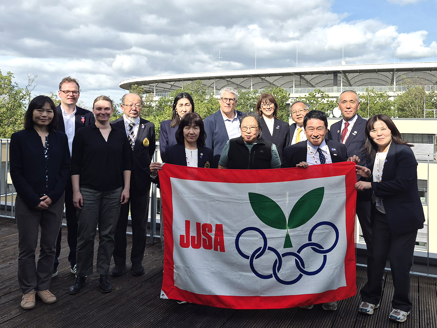 Eine Gruppe von Personen steht auf einer Terrasse vor einem Stadion. Sie halten eine Flagge mit dem Logo der JJSA und den olympischen Ringen. Die Szene zeigt eine Mischung aus formeller Kleidung und freundlichen Gesichtern, unter einem bewölkten Himmel.
