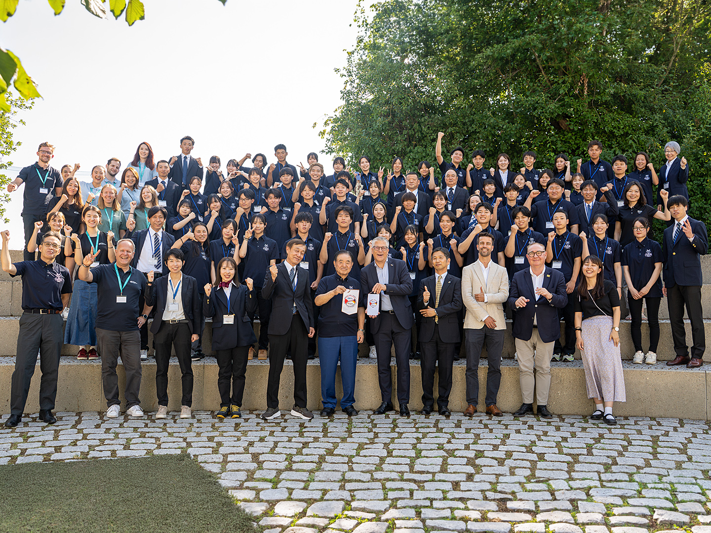 Eine große Gruppe von überwiegend japanischen Menschen steht auf einer Treppe im Freien, lächelt und posiert für ein Foto. Die Teilnehmenden tragen überwiegend schwarze T-Shirts. Im Hintergrund sind Bäume und ein klarer Himmel sichtbar.