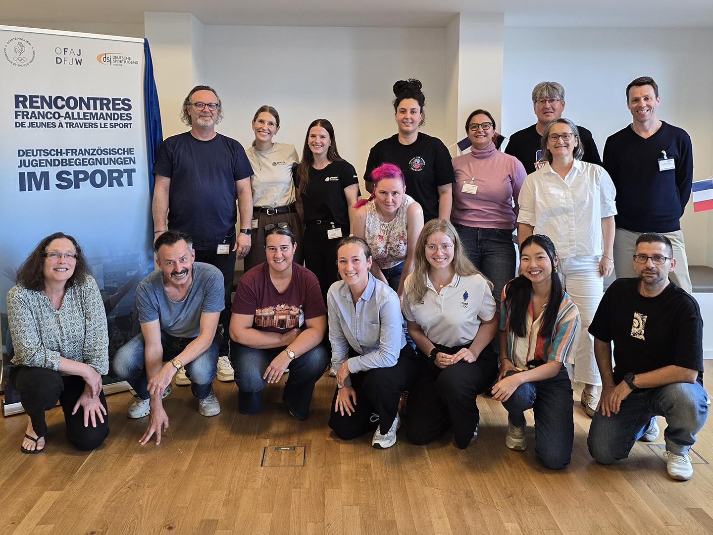Eine Gruppe von 16 Personen steht und sitzt in einem Raum, umgeben von einem Banner mit dem Text "RENCONTRES FRANCO-ALLEMANDES DE JEUNES TRAVAILLEURS DU SPORT". Die Teilnehmer sind vielfältig gekleidet und lächeln in die Kamera.