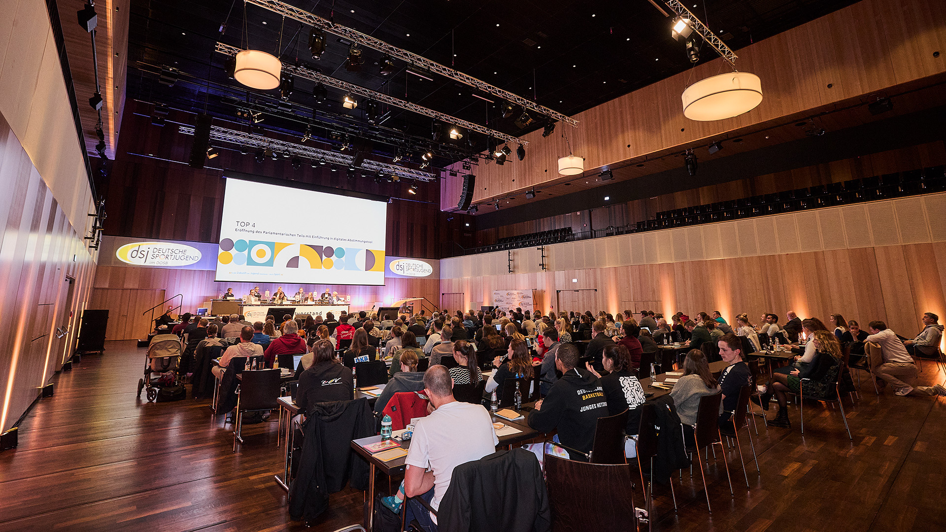 Großer Konferenzsaal mit zahlreichen Personen, die an Tischen sitzen und auf eine Bühne mit einer großen Leinwand blicken. Auf der Leinwand sind bunte grafische Elemente und der Schriftzug 'TOP 4' zu sehen. Die Bühne ist mit mehreren Personen besetzt, die an einem langen Tisch sitzen. Der Raum ist modern gestaltet mit Holzverkleidungen an den Wänden und großen runden Deckenleuchten. Die Veranstaltung findet im Rahmen des Hauptausschusses der Deutschen Sportjugend in Ingelheim am Rhein 2025 statt.