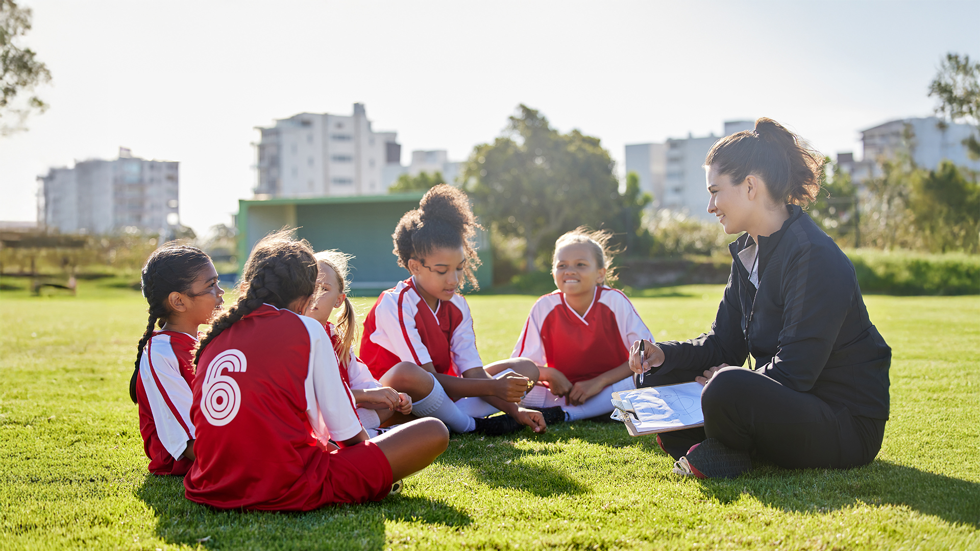 Eine Trainerin sitzt auf einer Wiese und bespricht mit einer Gruppe junger Mädchen in roten Sporttrikots eine Übungseinheit auf einem Sportplatz, im Hintergrund sind Grünflächen und Wohngebäude zu sehen.